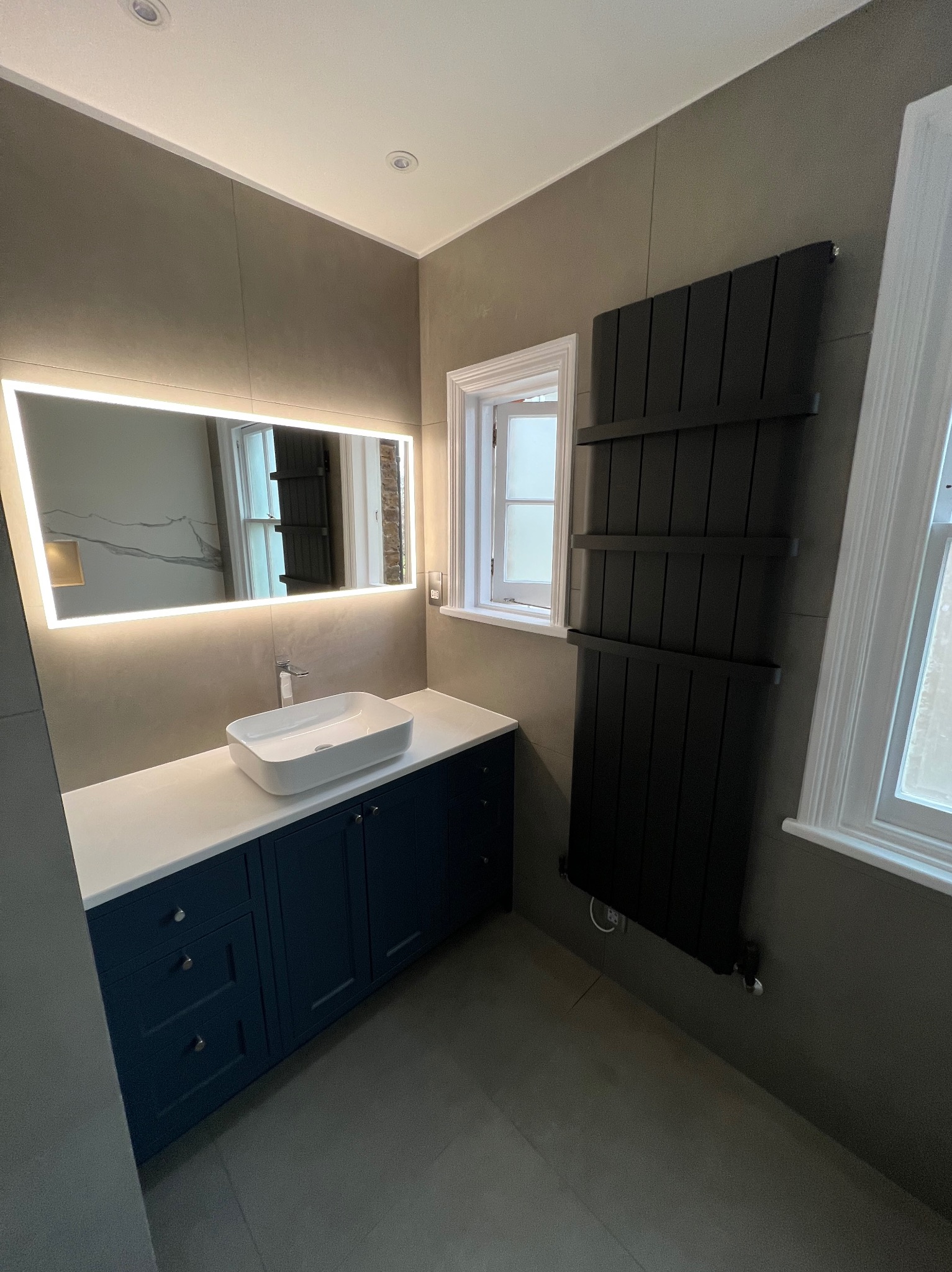 Nowoczesna łazienka z szarymi ścianami, podświetlanym lustrem nad białą umywalką nablatową na szafce w kolorze granatowym oraz czarnym grzejnikiem drabinkowym obok okna.