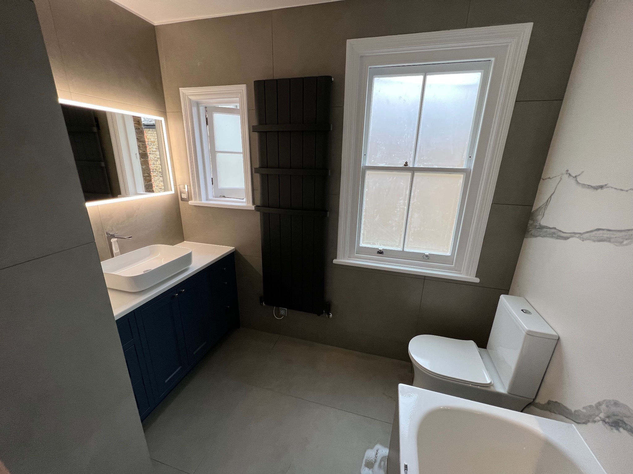 Nowoczesna łazienka w szaro-niebieskiej tonacji z podświetlanym lustrem, grzejnikiem drabinkowym i płytkami imitującymi marmur na jednej ze ścian.