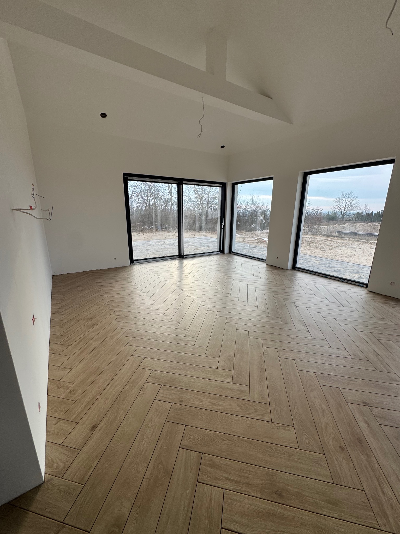 Salon w nowym domu z ułożoną podłogą z paneli w jodełkę, widoczne duże okna z czarnymi ramami i fragmenty instalacji elektrycznej.