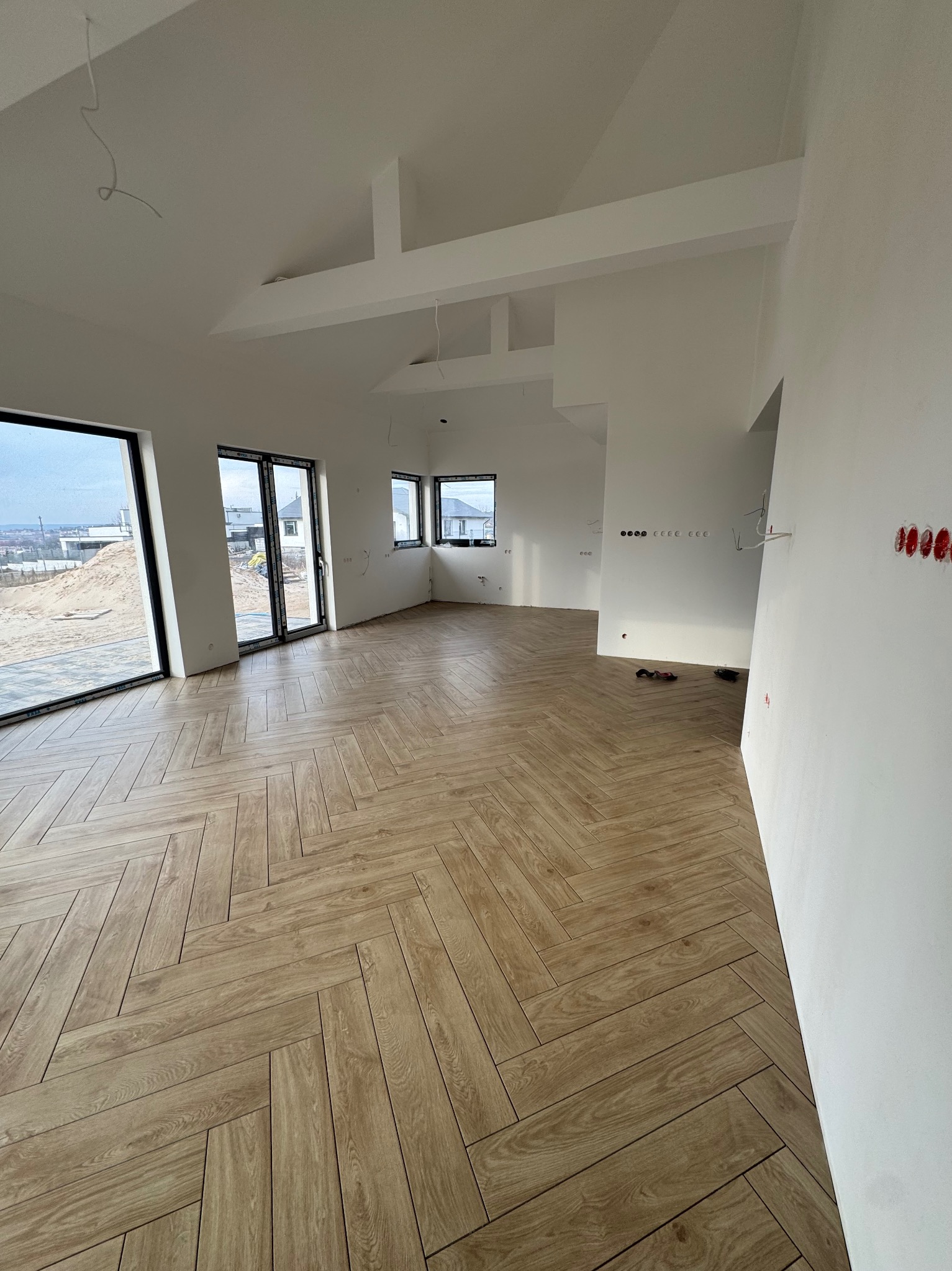 Jasne panele ułożone we wzór jodełki w przestronnym, surowym wnętrzu z dużymi oknami i białymi ścianami, widoczne gniazdka elektryczne i wystające kable.