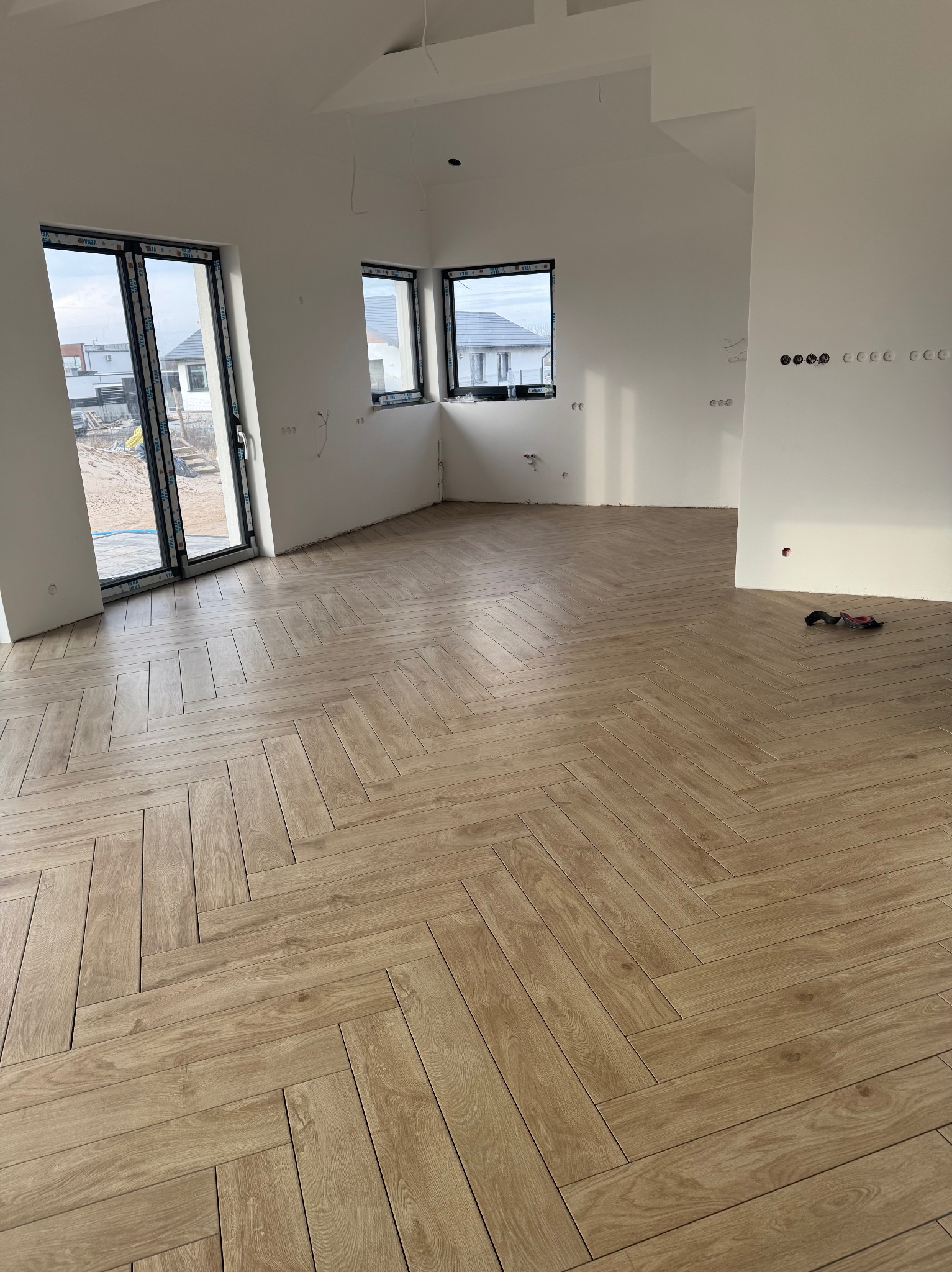 Ułożona w jodełkę podłoga z paneli drewnopodobnych w jasnym, przestronnym wnętrzu z dużymi oknami; widoczne narzędzia glazurnicze w rogu.