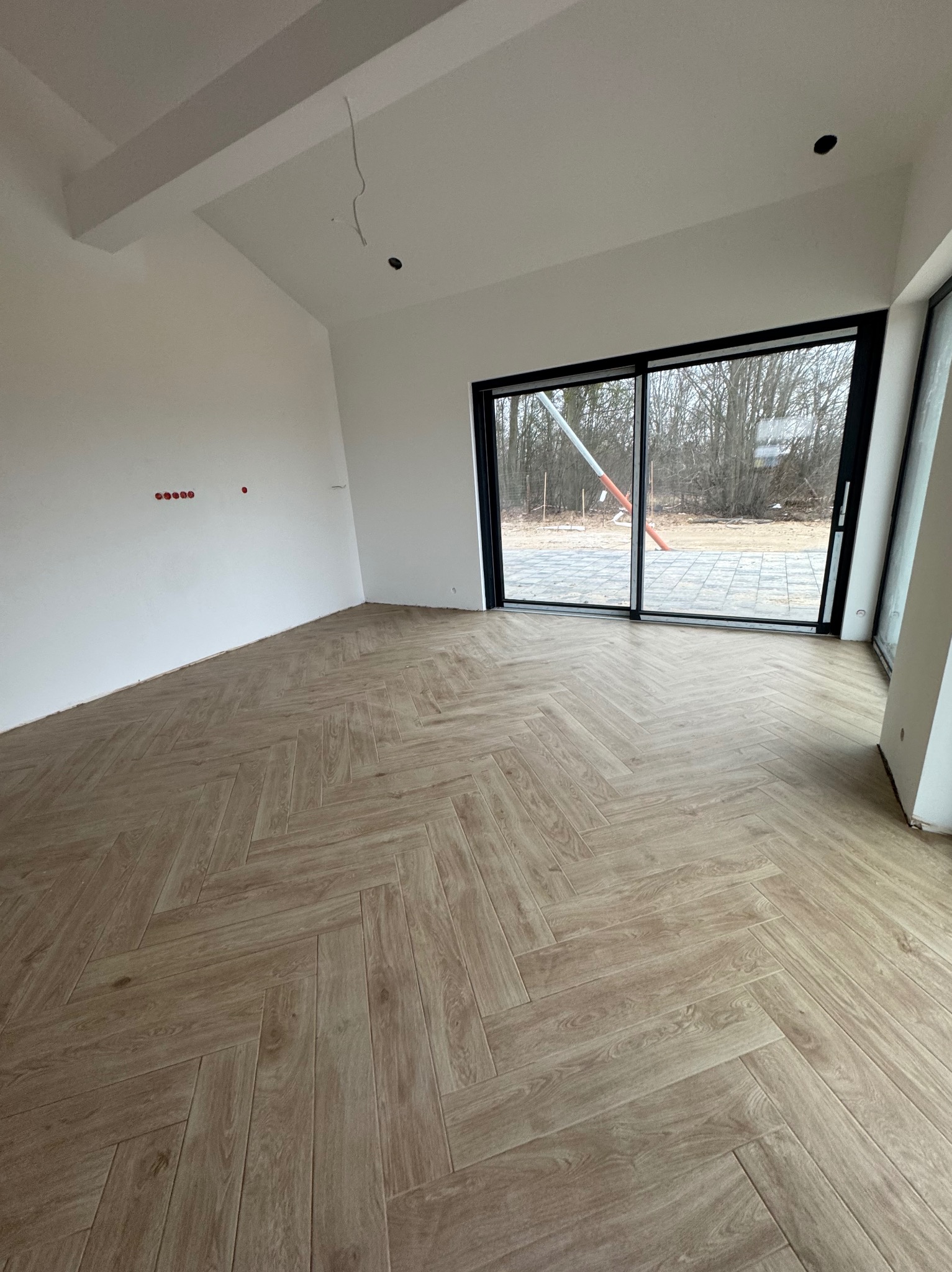 Jasne panele ułożone w jodełkę w przestronnym, surowym wnętrzu z dużymi przesuwnymi oknami i widocznymi instalacjami elektrycznymi.