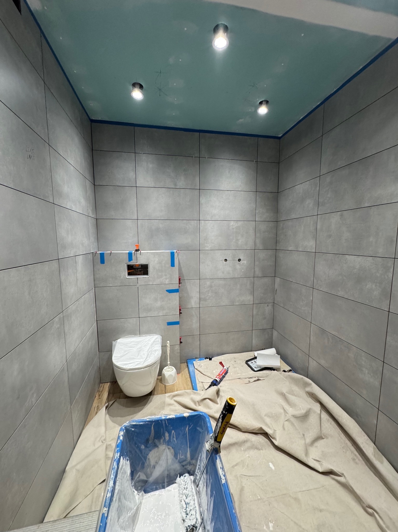 Łazienka w trakcie prac w Lubichowie: szare płytki, podwieszana toaleta z osprzętem, malowanie sufitu na niebiesko, narzędzia malarskie.