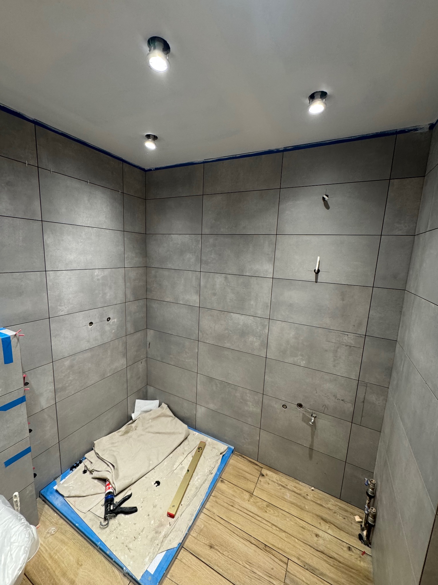Łazienka w trakcie remontu: szare płytki na ścianach, drewnopodobna podłoga, widoczne narzędzia glazurnicze i przygotowane punkty oświetleniowe na suficie.