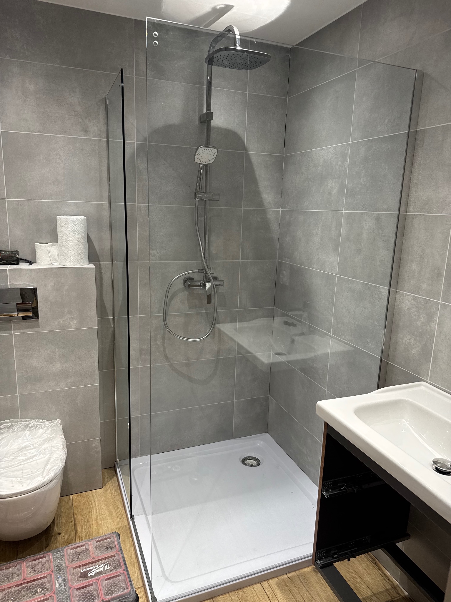 Nowoczesna kabina prysznicowa ze szklaną ścianką i deszczownicą, szare płytki na ścianach. Widoczna umywalka z szafką i toaleta z podwieszanym systemem spłukiwania.