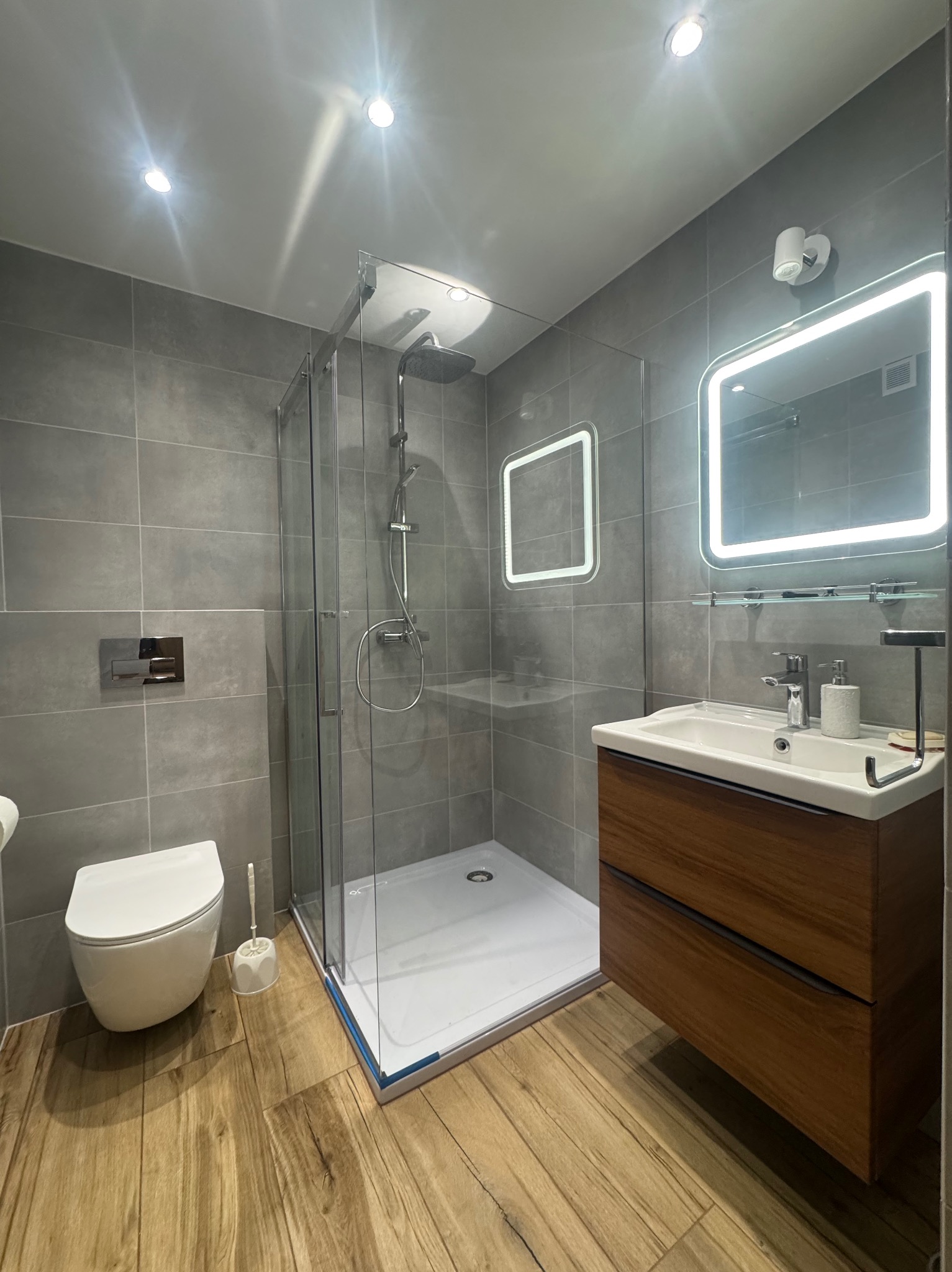 Nowoczesna łazienka w szarościach z drewnianą szafką pod umywalką i kabiną prysznicową z deszczownicą. Lustro z podświetleniem LED.