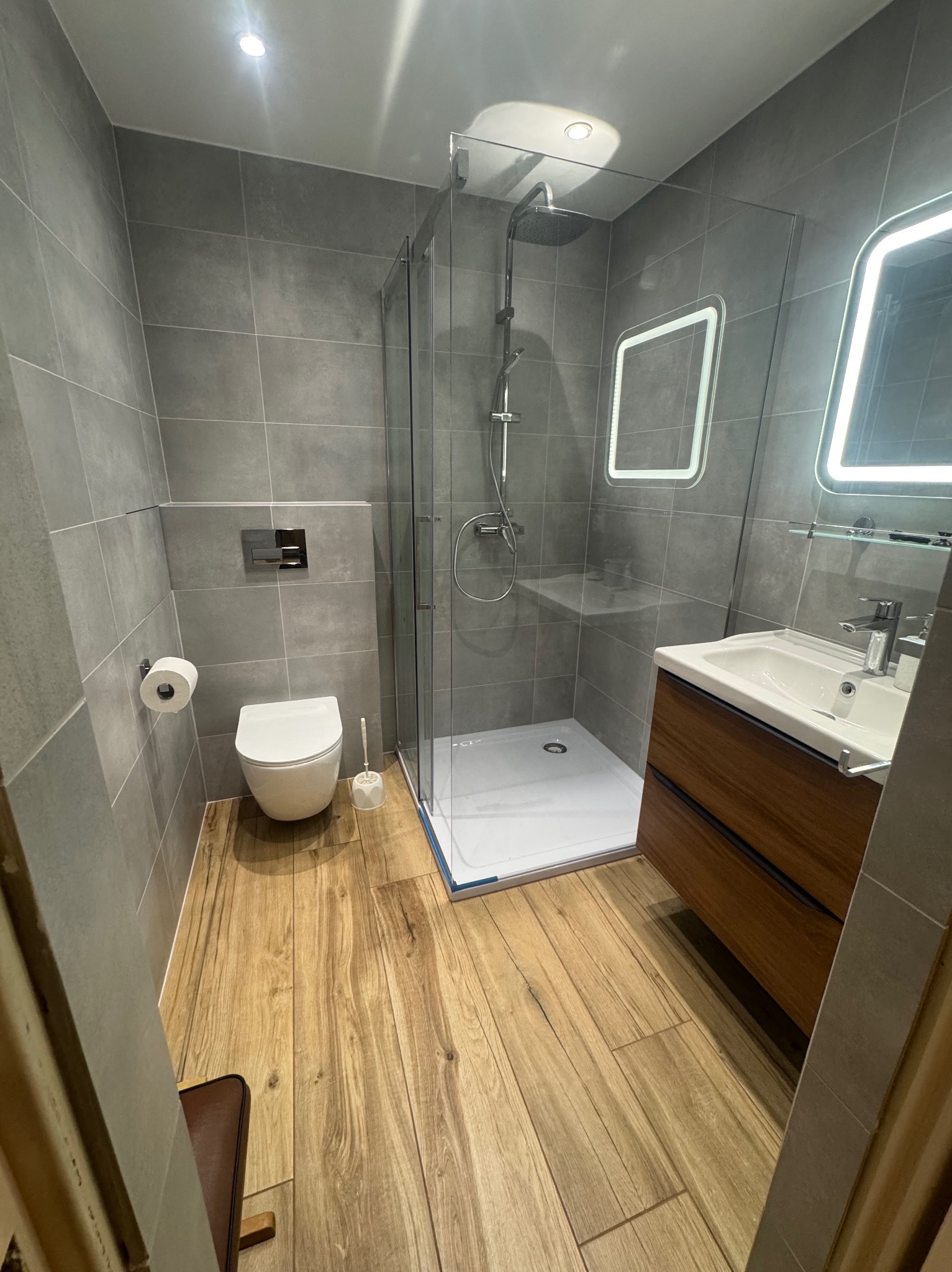 Nowoczesna łazienka po remoncie: szare płytki ścienne, drewnopodobna podłoga, kabina prysznicowa ze szkła, podwieszana toaleta i umywalka z szafką z ciemnego drewna.