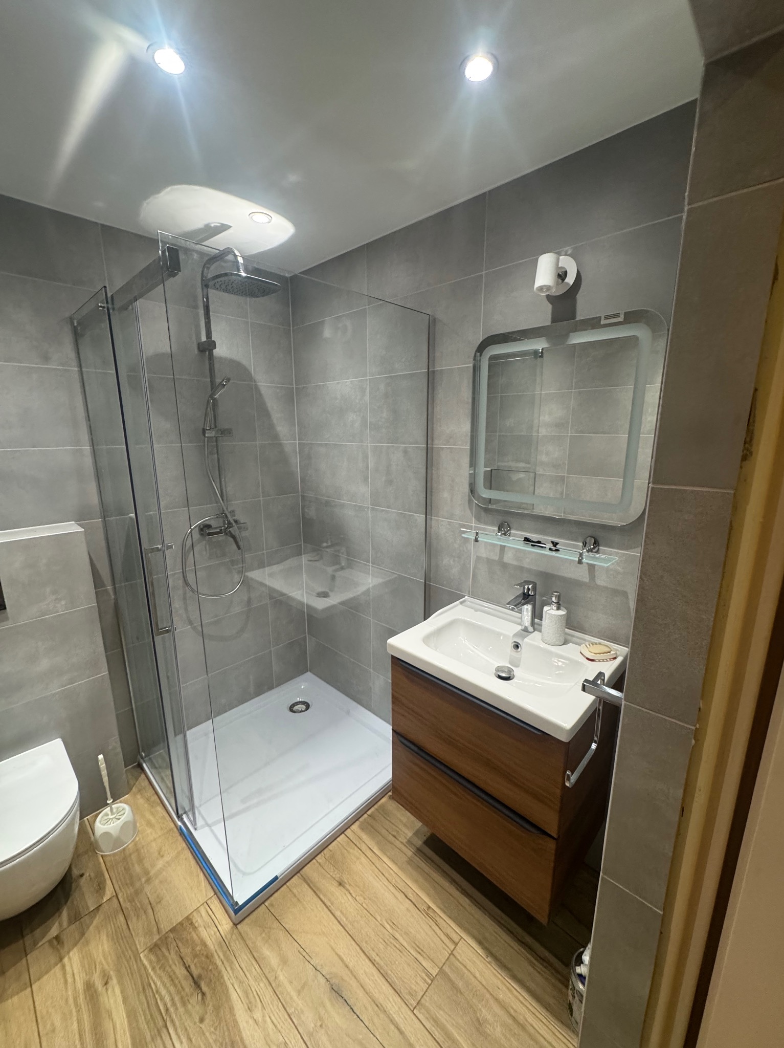 Nowoczesna łazienka w szarości z drewnianymi akcentami: kabina prysznicowa ze szklanymi ścianami, umywalka z szafką i lustro z oświetleniem LED.