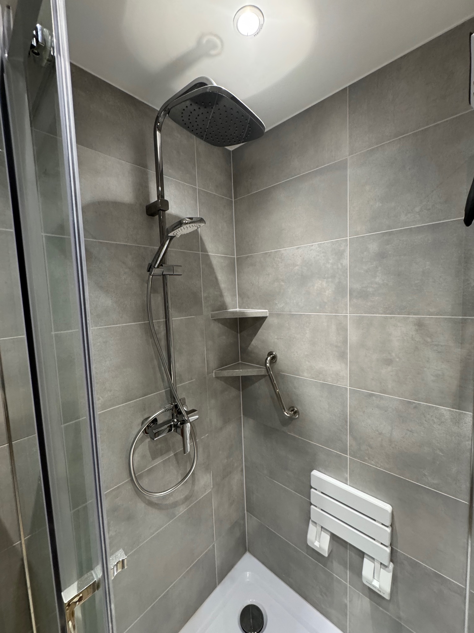 Nowoczesna kabina prysznicowa z szarymi płytkami, deszczownicą, półkami narożnymi i składanym siedziskiem. Minimalistyczny design, jasne oświetlenie.