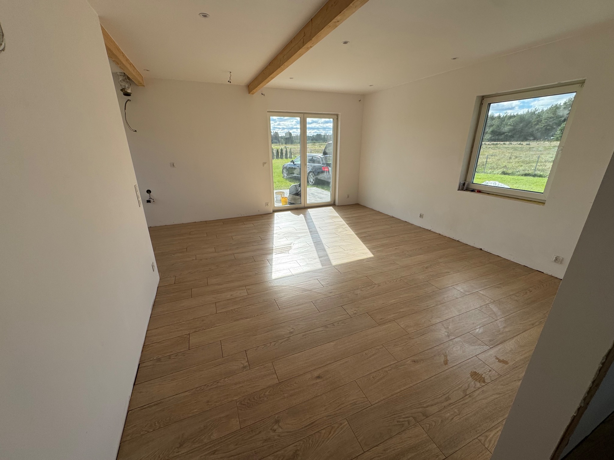 Jasne, przestronne wnętrze z drewnopodobną podłogą z płytek, widok na zewnątrz przez okno i drzwi balkonowe. Naturalne światło podkreśla ciepły odcień podłogi.