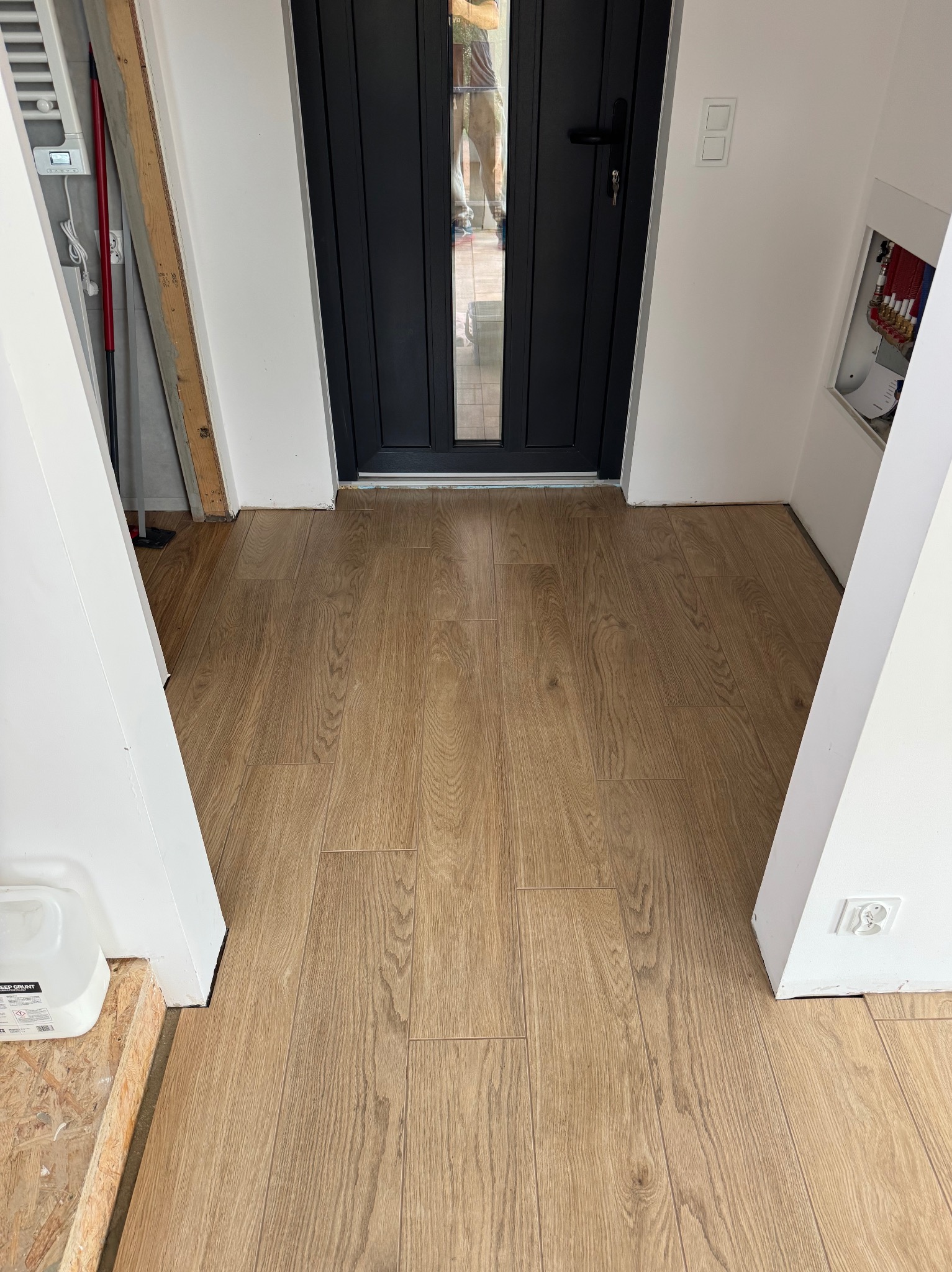 Ułożone panele podłogowe w przedpokoju, widok z góry. Jasne panele imitujące drewno, czarne drzwi wejściowe, białe ściany. Widoczny fragment instalacji elektrycznej.