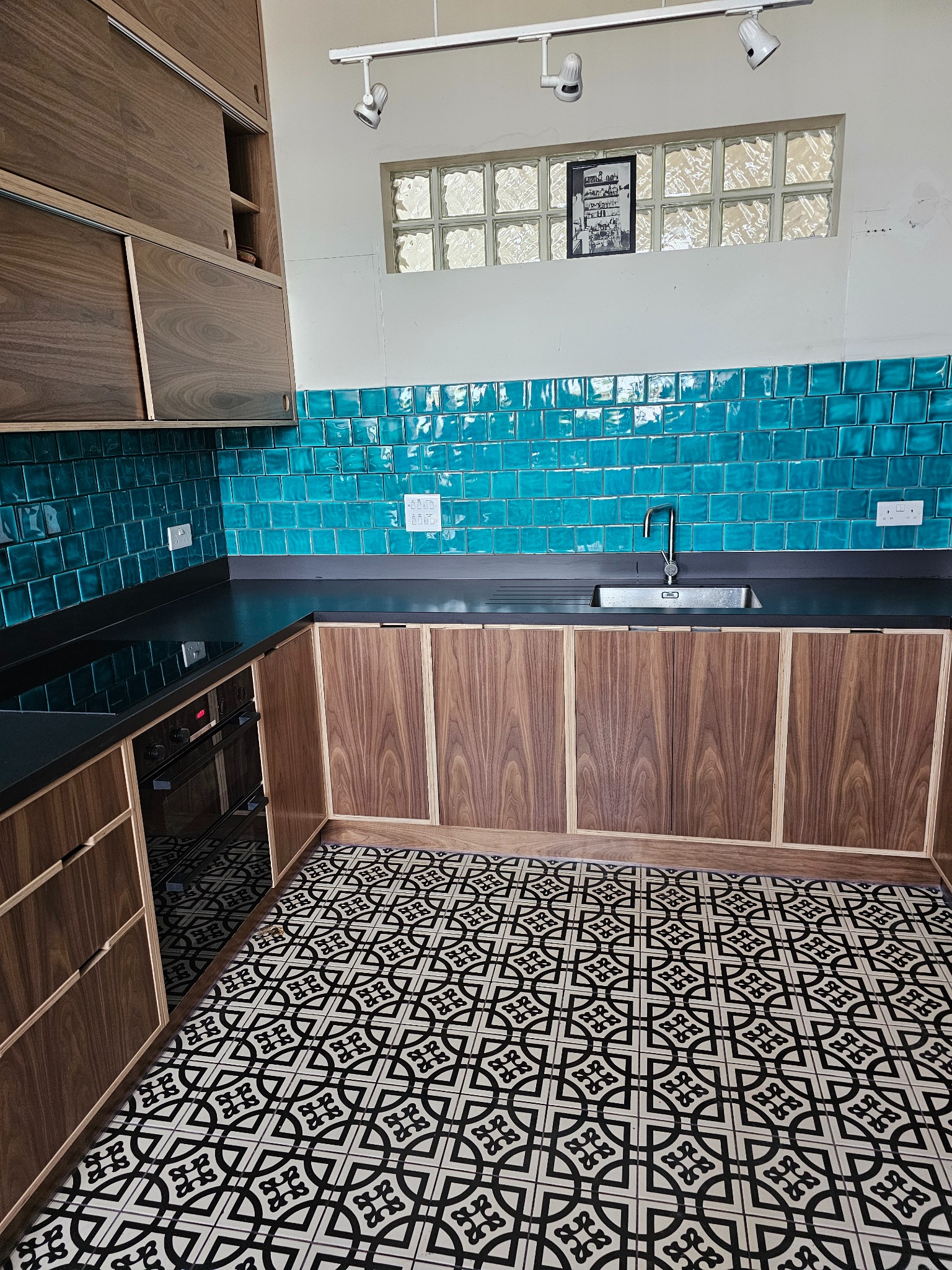 Nowoczesna kuchnia z turkusowymi płytkami ściennymi, drewnianymi szafkami z forniru orzechowego i wzorzystą czarno-białą podłogą, oświetlona reflektorami sufitowymi.