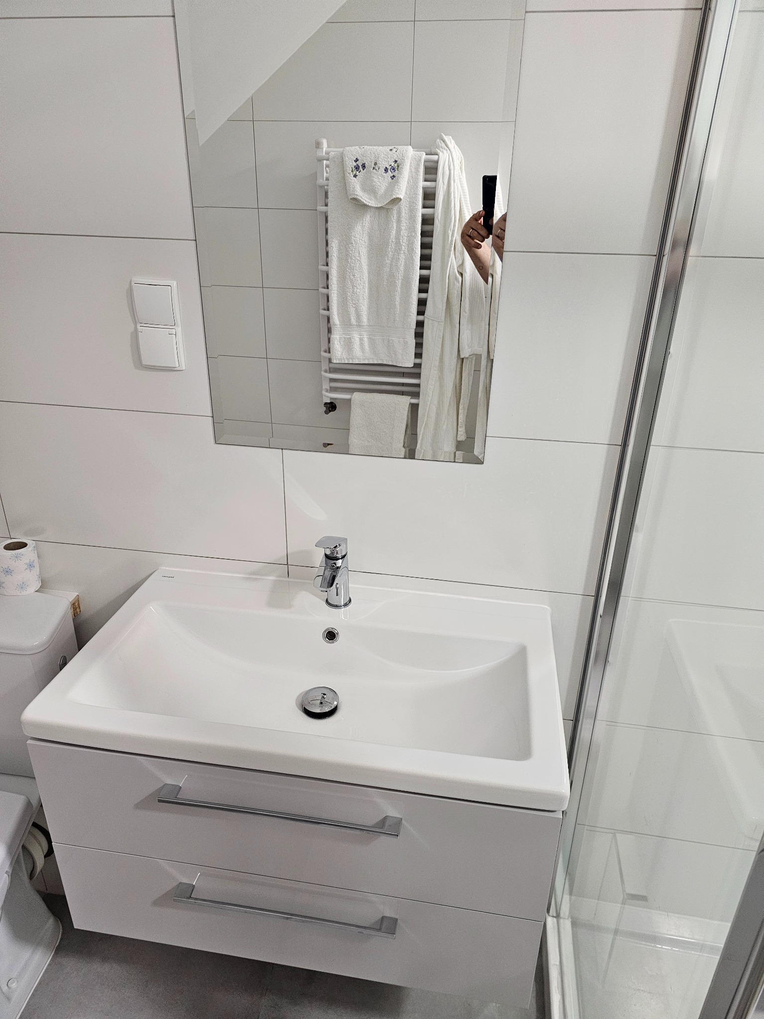 Biała umywalka z szafką i chromowaną baterią w nowoczesnej łazience z białymi płytkami, widoczne odbicie osoby robiącej zdjęcie w lustrze, ręczniki na grzejniku drabinkowym w tle.
