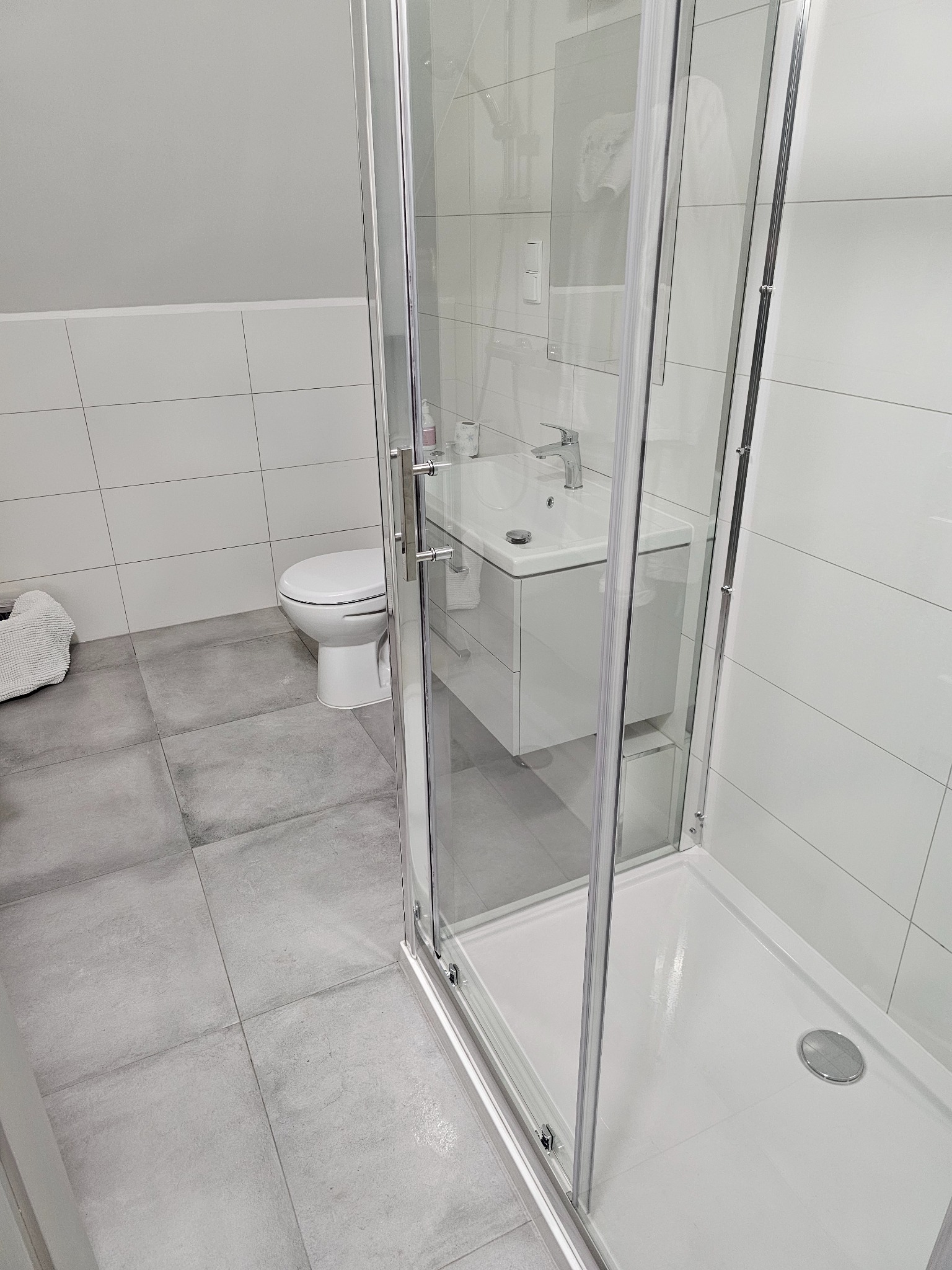 Nowoczesna łazienka z kabiną prysznicową ze szklanymi drzwiami, białą umywalką z szafką i toaletą w tle, wykończona szarymi płytkami podłogowymi i białymi płytkami ściennymi.