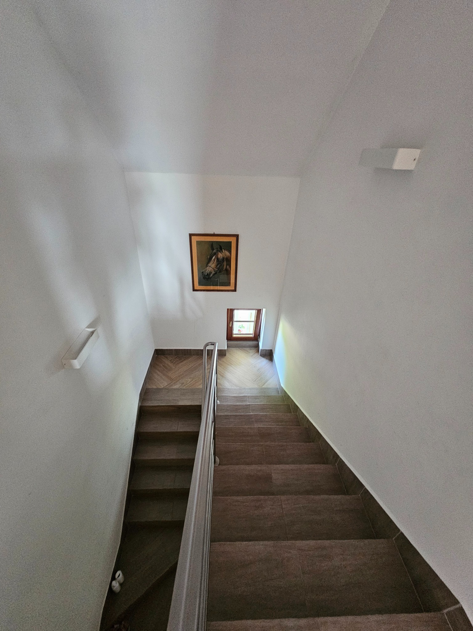 Klatka schodowa wykończona brązowymi płytkami imitującymi drewno, z metalową barierką, pomalowanymi na biało ścianami, obrazem konia i małym oknem na końcu biegu schodów.