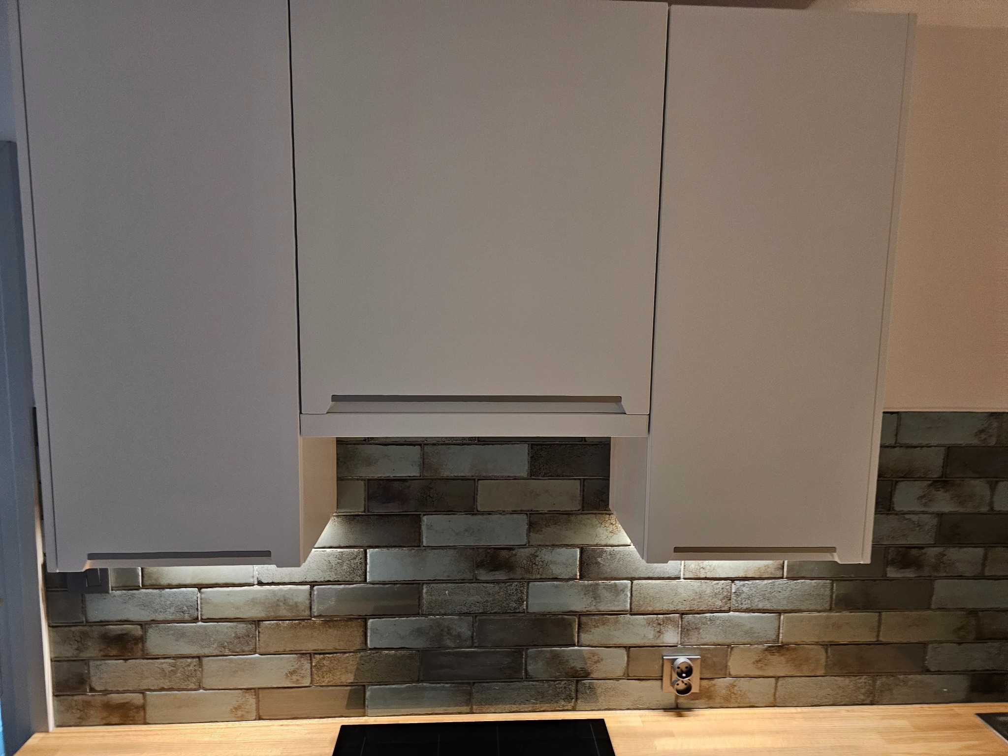 Białe szafki kuchenne wiszące nad blatem z drewnianym wykończeniem i dekoracyjną ścianą z cegieł oświetloną od dołu.