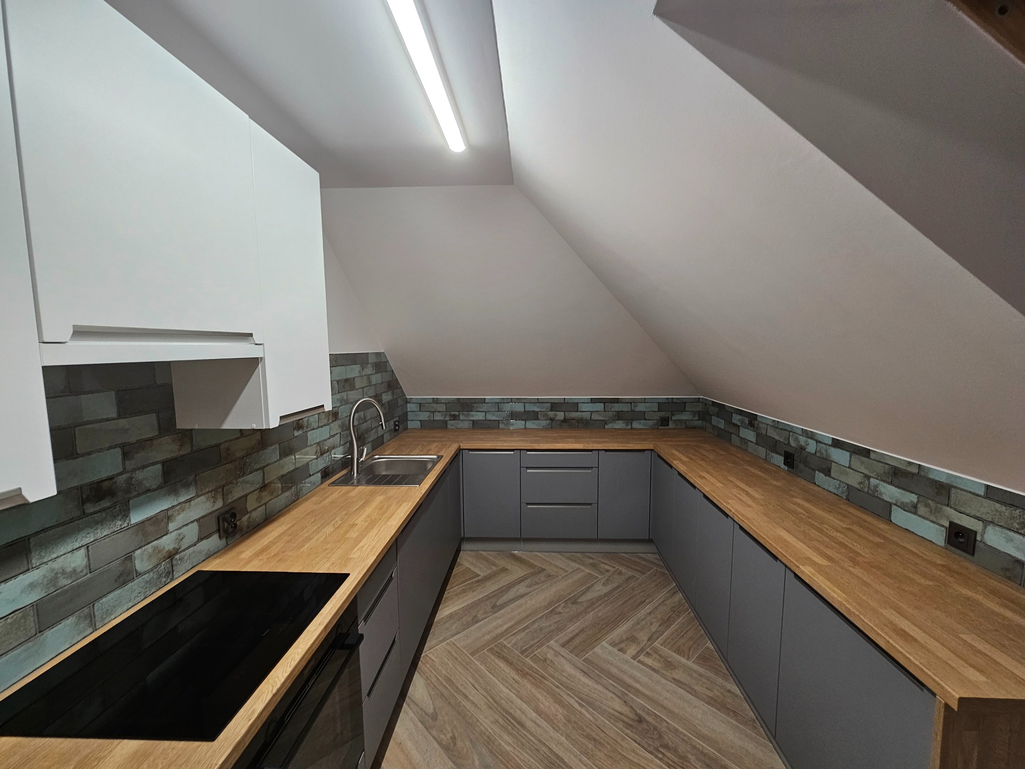 Kuchnia w szarych tonach z drewnianym blatem i podłogą ułożoną w jodełkę, oświetlona lampą sufitową, w pomieszczeniu ze skośnym sufitem.