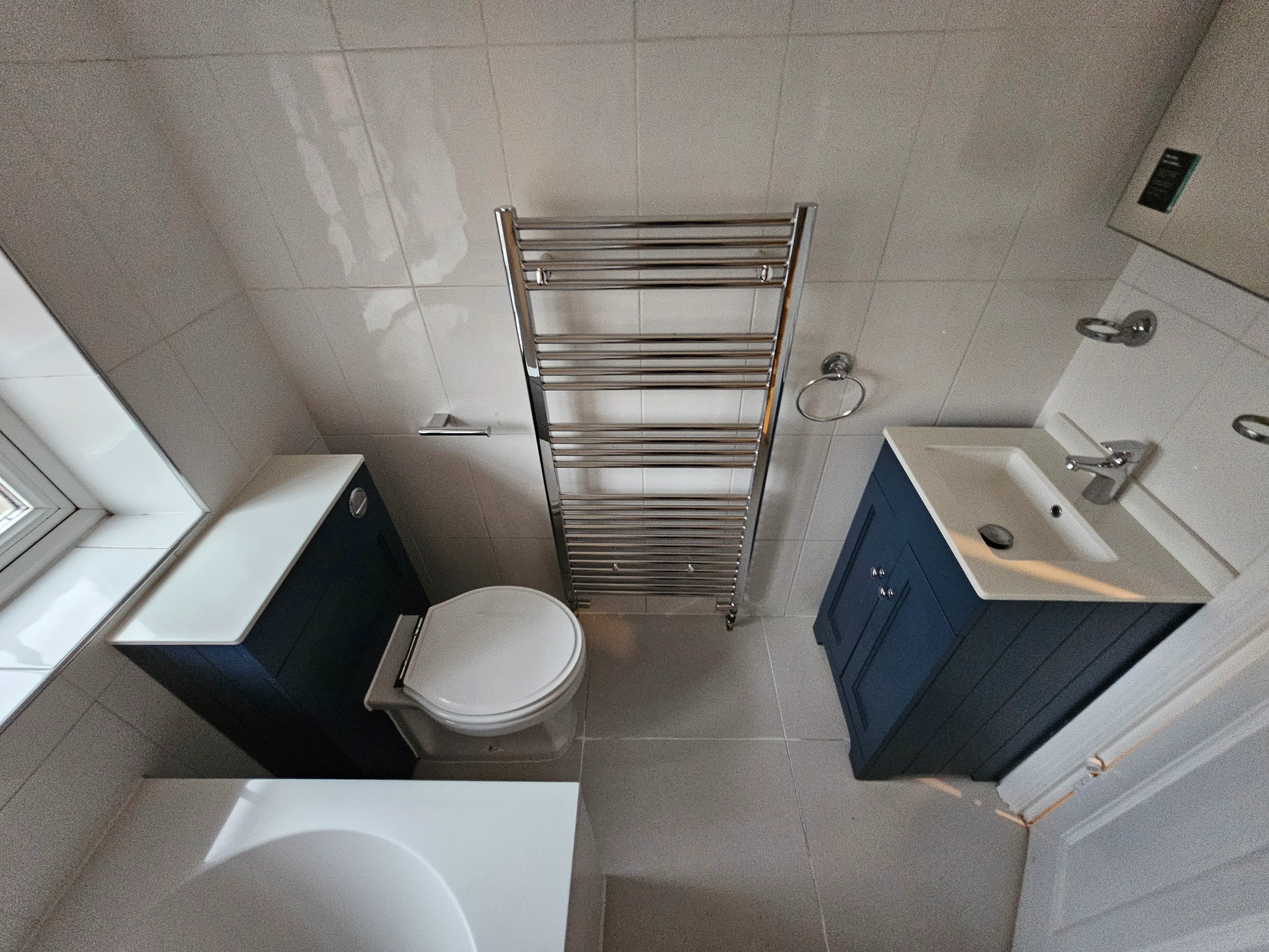 Widok z góry na wyremontowaną łazienkę z białymi płytkami, z toaletą obok szafki pod umywalką w kolorze granatowym, grzejnik drabinkowy w chromie i fragment wanny.