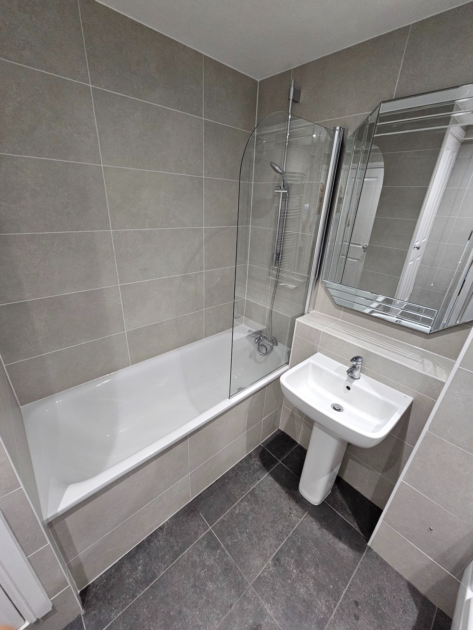 Nowoczesna łazienka po remoncie: szare płytki na ścianach i podłodze, biała wanna z parawanem nawannowym, umywalka na postumencie i lustro z fazowanymi krawędziami.