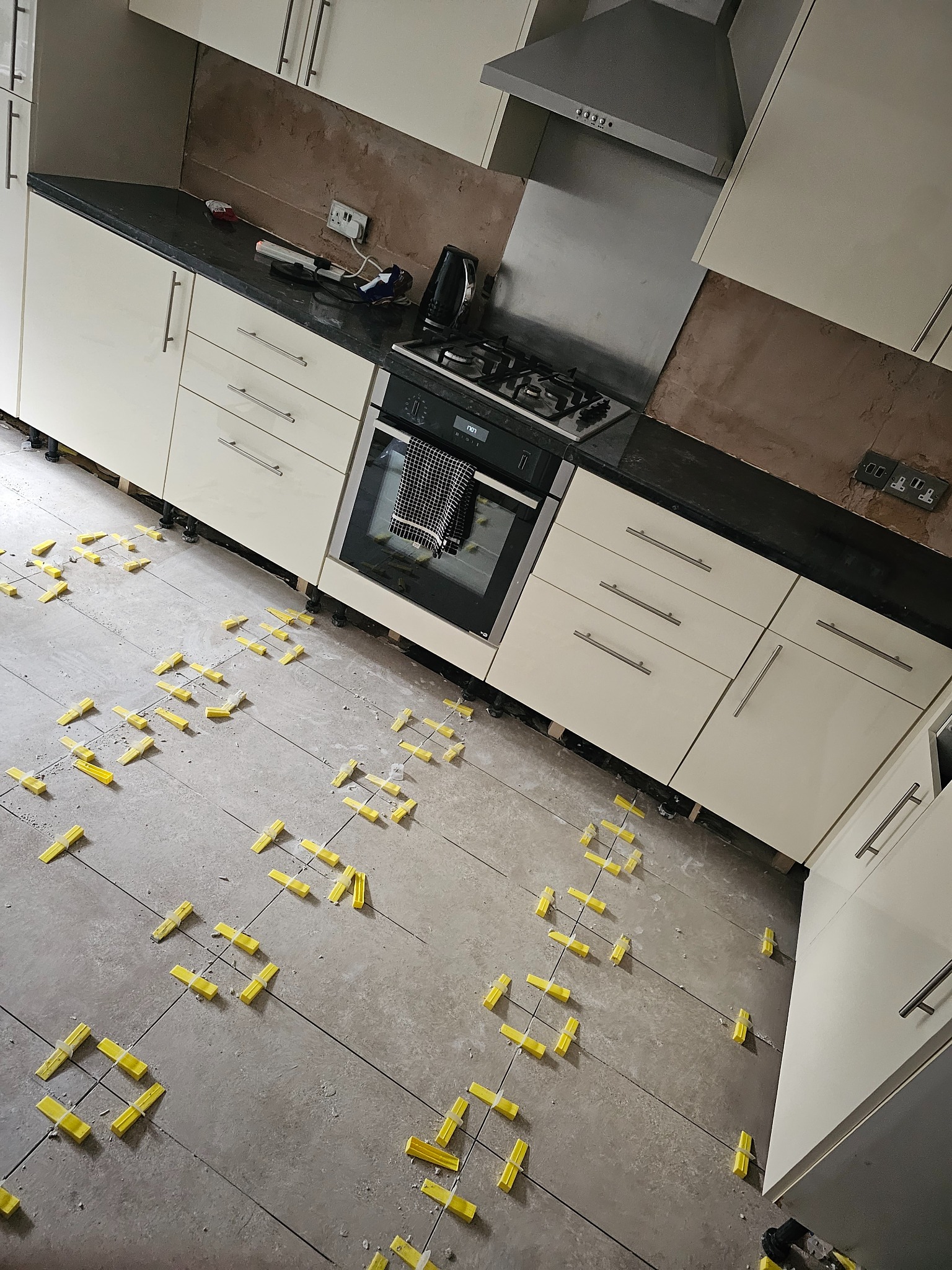 Układanie szarych płytek podłogowych w kuchni z użyciem żółtych klinów poziomujących, widoczne kremowe szafki kuchenne i sprzęt AGD