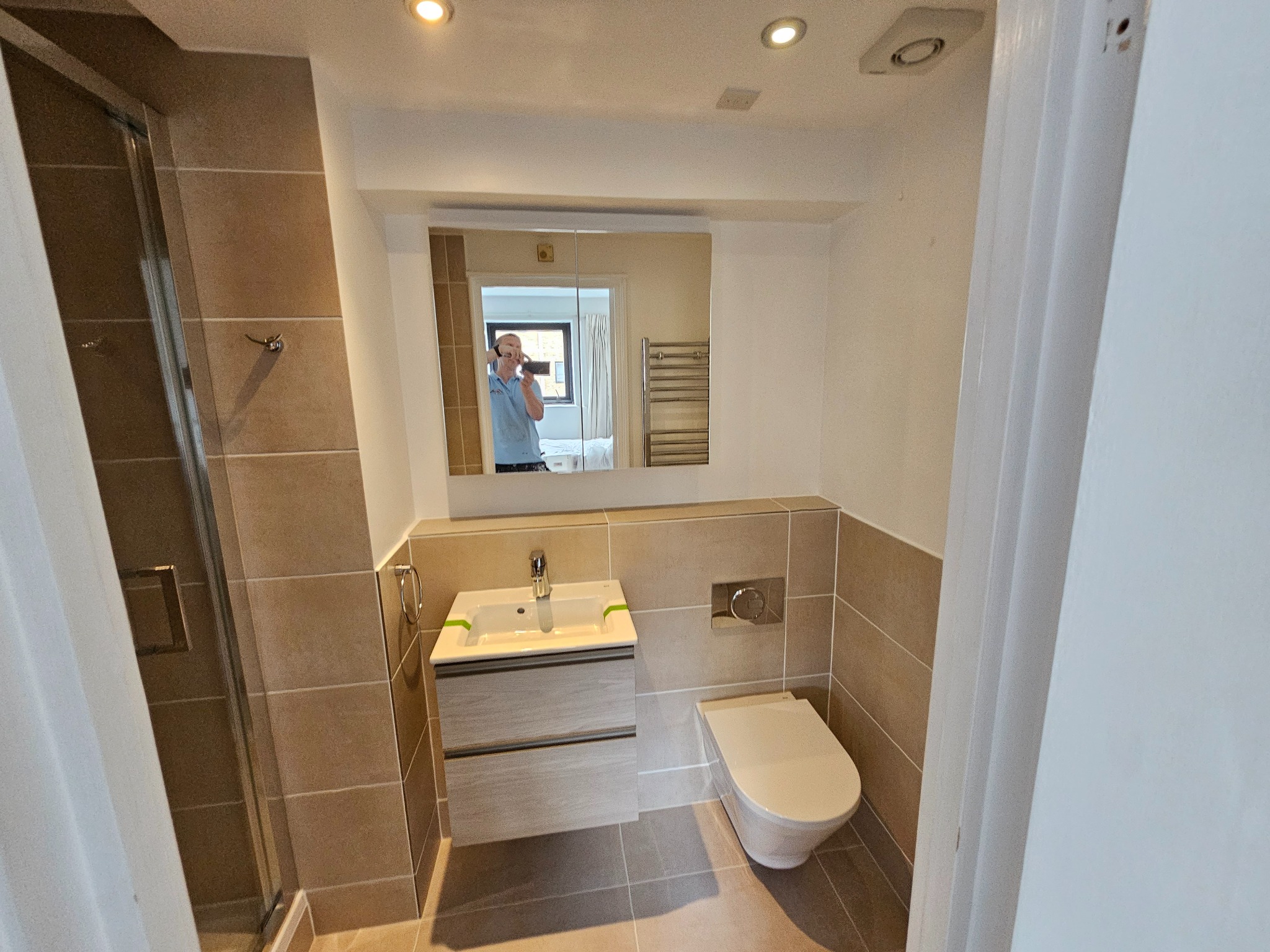 Nowoczesna łazienka z beżowymi płytkami, kabiną prysznicową, podwieszaną szafką z umywalką i lustrem, w którym odbija się osoba robiąca zdjęcie.