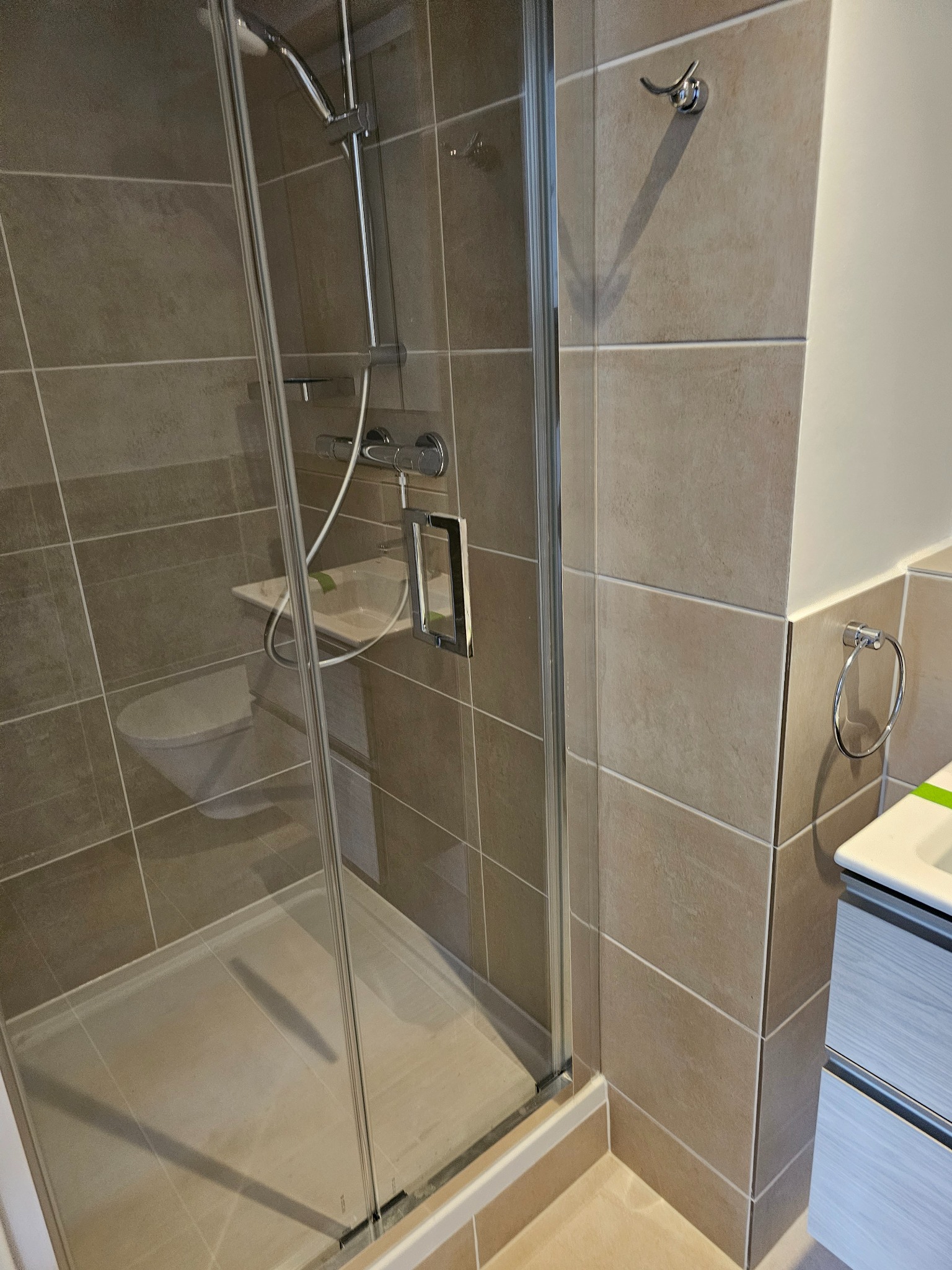 Beżowa łazienka z kabiną prysznicową typu walk-in, widoczna toaleta w tle i szafka pod umywalką z szufladami o wzorze imitującym drewno.