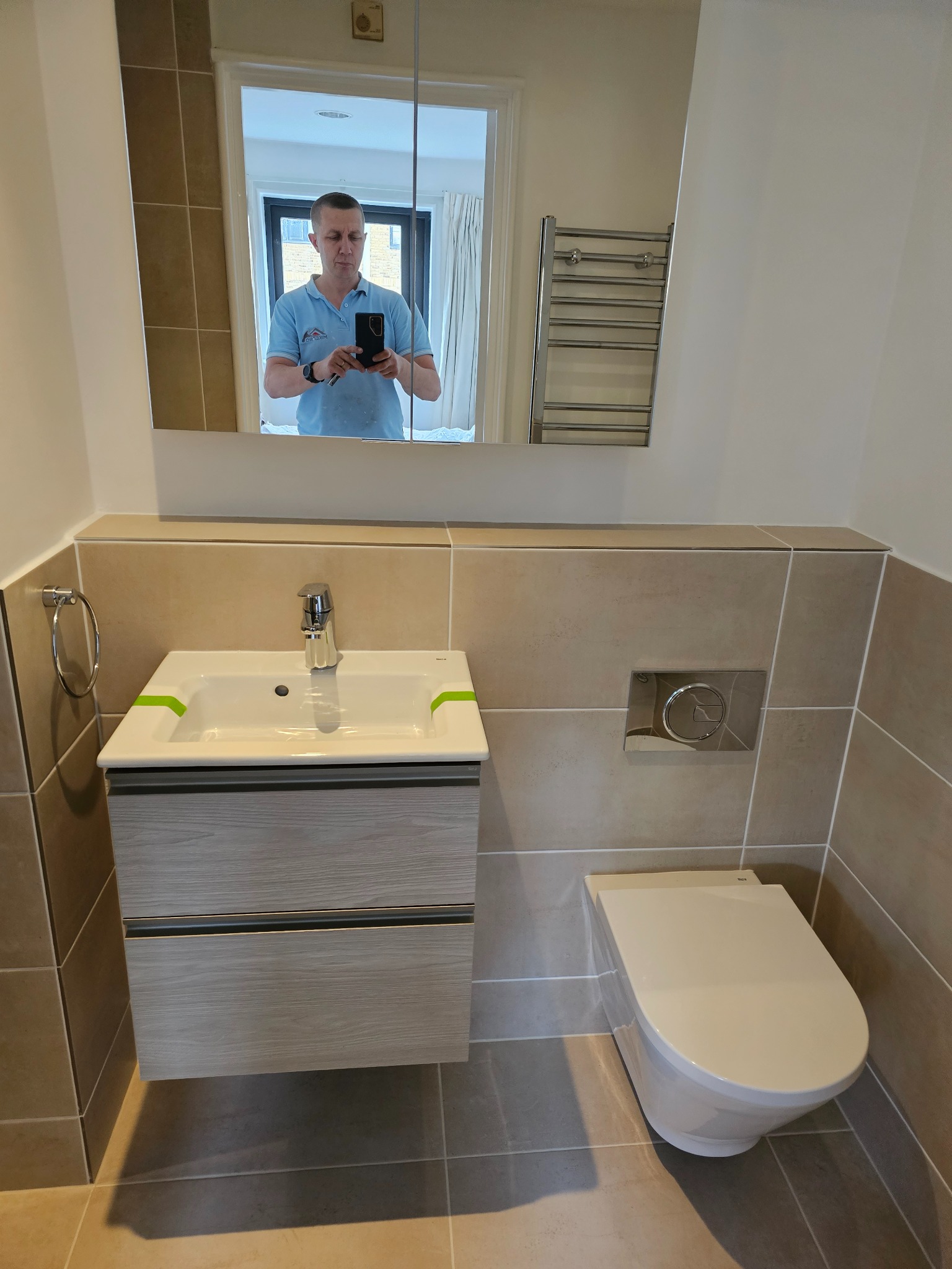 Nowoczesna łazienka z beżowymi płytkami, wiszącą toaletą i umywalką z szafką w kolorze jasnego drewna; w lustrze odbija się mężczyzna robiący zdjęcie.