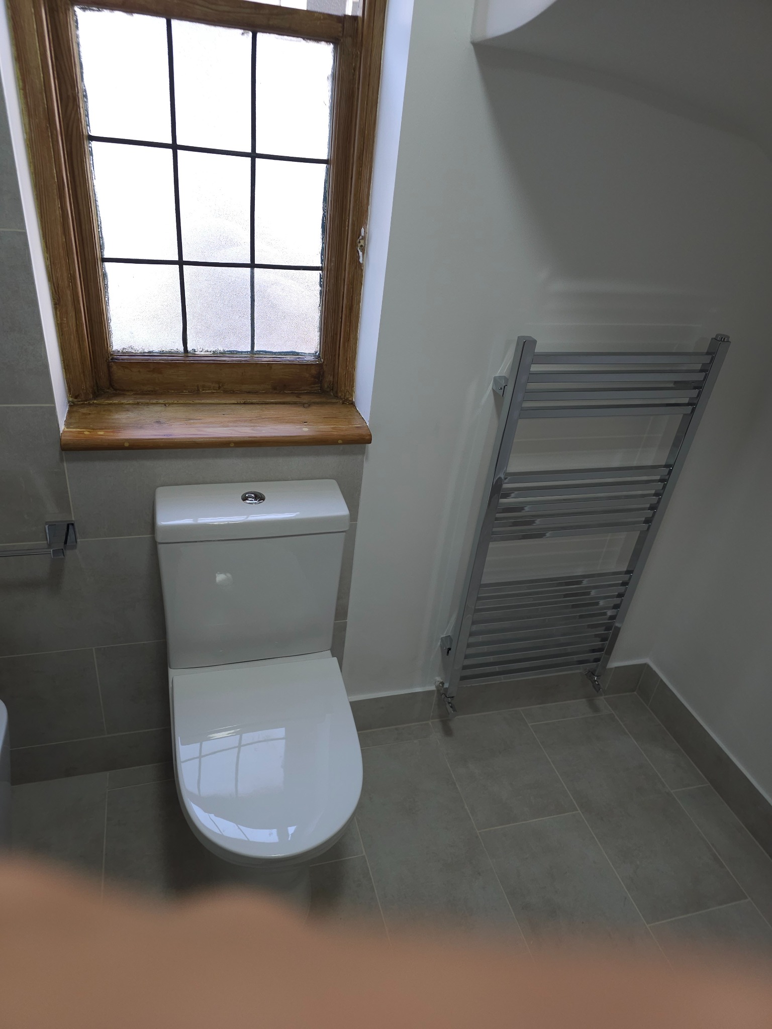 Nowoczesna łazienka z wiszącą toaletą, grzejnikiem drabinkowym i oknem z drewnianą ramą, wykończona szarą płytką.