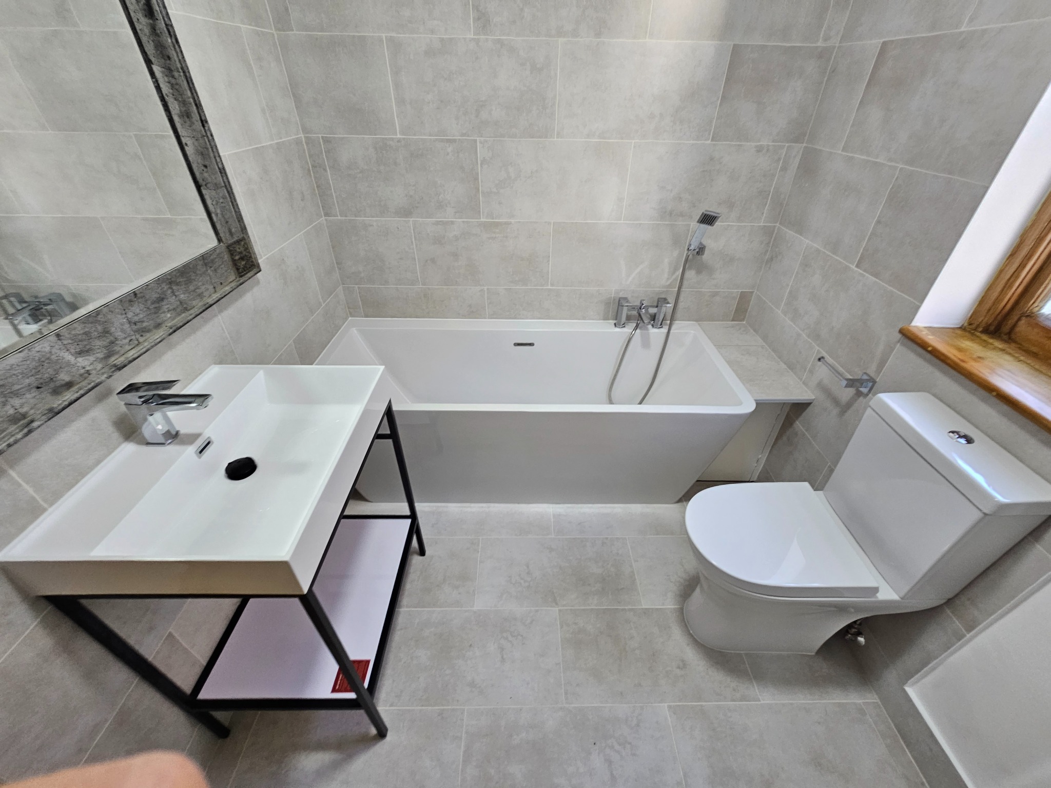 Nowoczesna łazienka z szarymi płytkami, białą wanną prostokątną, umywalką na czarnym stelażu z białą półką i toaletą kompaktową, wykończona drewnianym oknem.