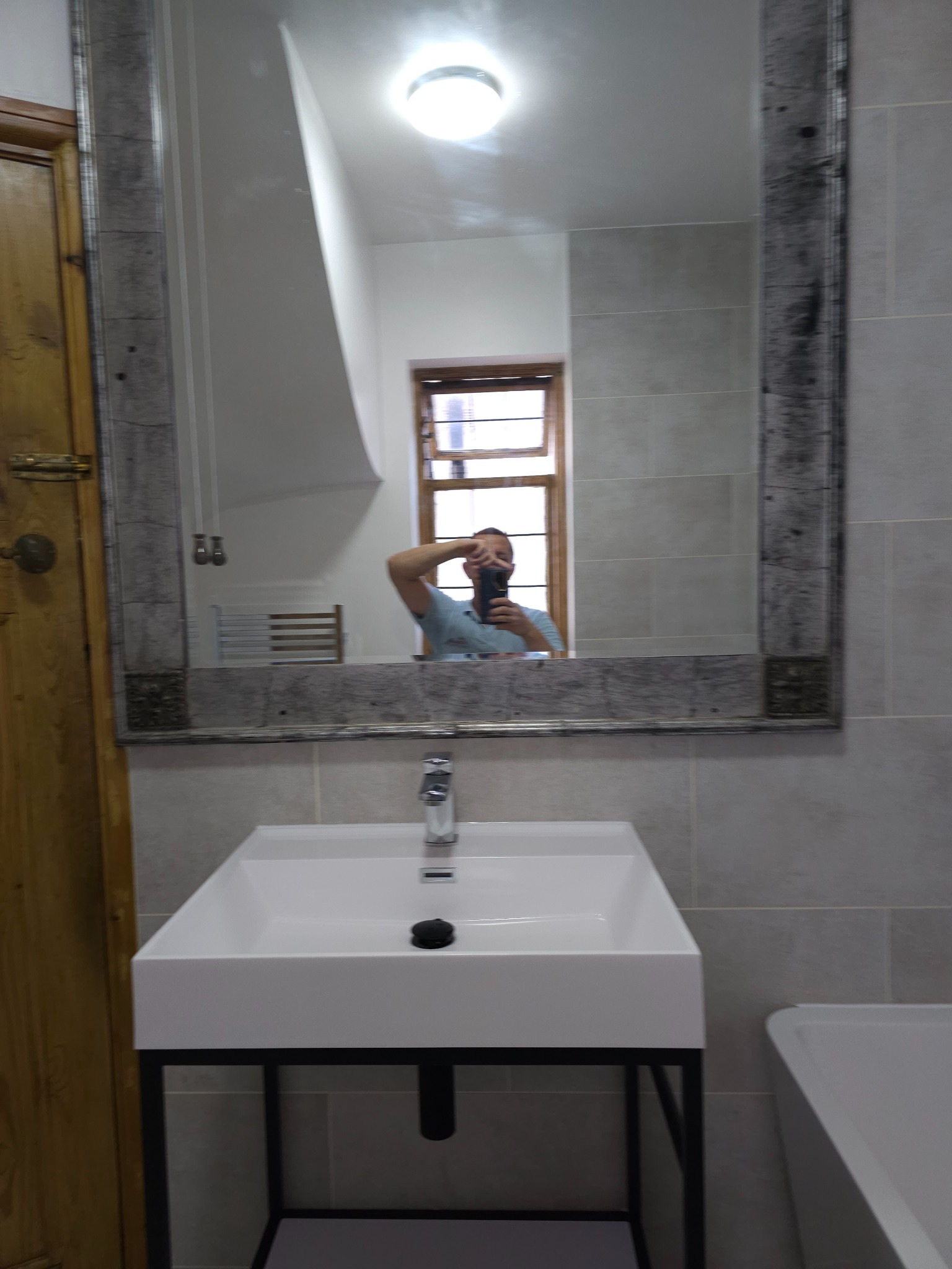 Nowoczesna biała umywalka nablatowa z czarnym syfonem, chromowana bateria, lustro w drewnianej ramie z efektem postarzenia, szare płytki na ścianie, odbicie fotografa w lustrze.