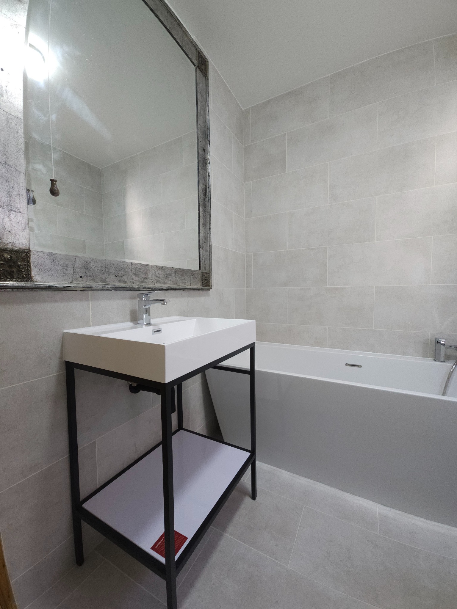 Nowoczesna łazienka w szarościach z wanną i umywalką na metalowym stelażu, lustro w ozdobnej ramie.