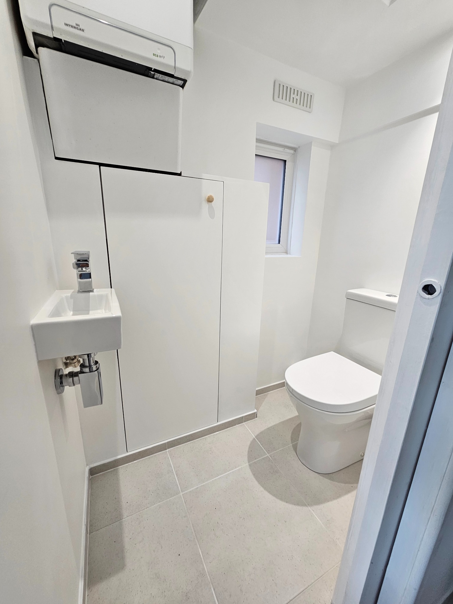 Nowoczesna, biała łazienka z wiszącą umywalką, zabudową meblową pod sufitem i toaletą. Widoczna kratka wentylacyjna i płytki podłogowe w odcieniach szarości.