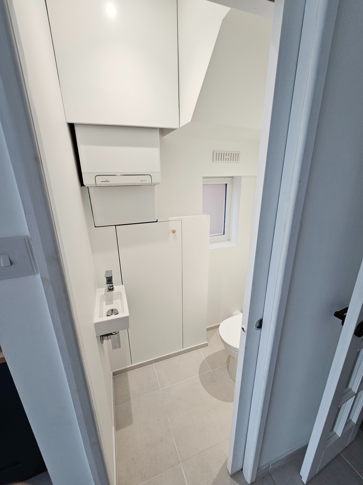 Mała, nowocześnie urządzona toaleta z wiszącą szafką, umywalką i grzejnikiem, wykończona w bieli i szarości, widoczna zza otwartych drzwi.