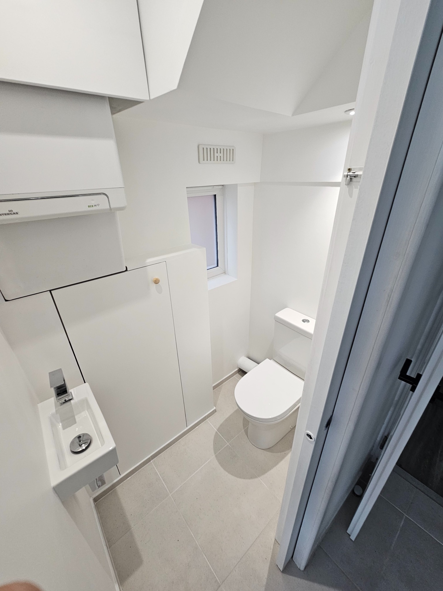 Mała, minimalistyczna toaleta z wiszącą umywalką, zabudową szafek pod sufitem i płytkami podłogowymi w odcieniach szarości, widziana zza częściowo otwartych drzwi.