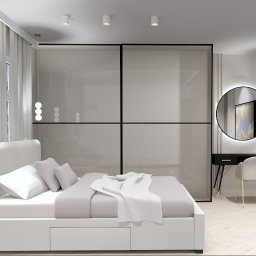 Trans - Hurt Sp. J. - Nowoczesna sypialnia w bieli i szarościach z łóżkiem z szufladami, przesuwną szafą z czarnymi ramami, okrągłym lustrem z podświetleniem LED nad czarną toaletką i parkietem w jodełkę.