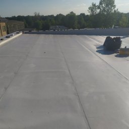 Alex Nikitin - Jasnoszara, płaska powierzchnia dachu z widocznymi łączeniami papy bitumicznej, na której leżą czarne worki na śmieci i żółta skrzynka narzędziowa. W tle widoczny budynek i drzewa, a na dachu stoi...