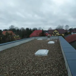 Płaski dach pokryty drobnym żwirem z trzema świetlikami i elementami wentylacyjnymi, widok z góry na tle zabudowy jednorodzinnej w pochmurny dzień.