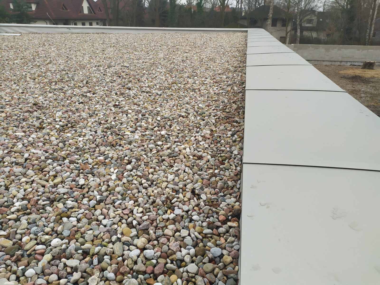 Płaski dach pokryty warstwą okrągłych, różnokolorowych kamieni, z widocznymi szarymi panelami na krawędziach i budynkami w tle.