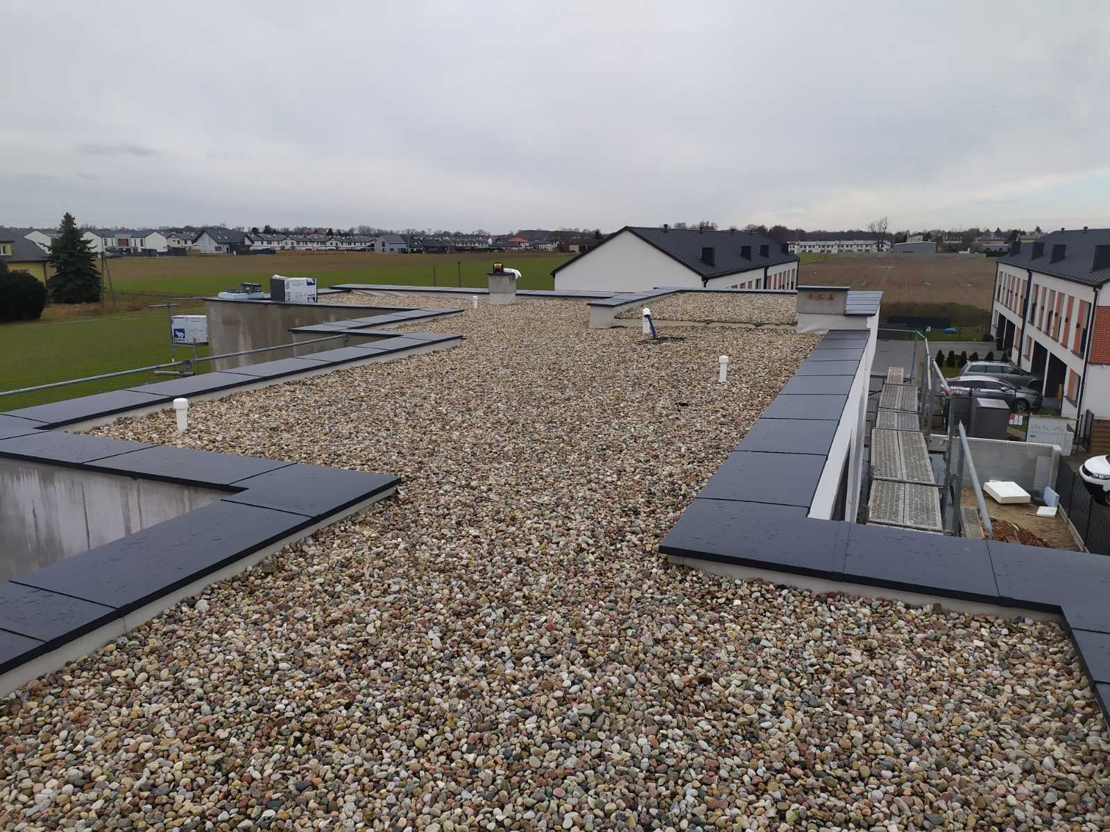 Widok z góry na płaski dach pokryty drobnymi kamykami, z widocznymi elementami wykończeniowymi w kolorze grafitowym i instalacjami wentylacyjnymi, na tle zabudowy mieszkalnej i pól uprawnych...