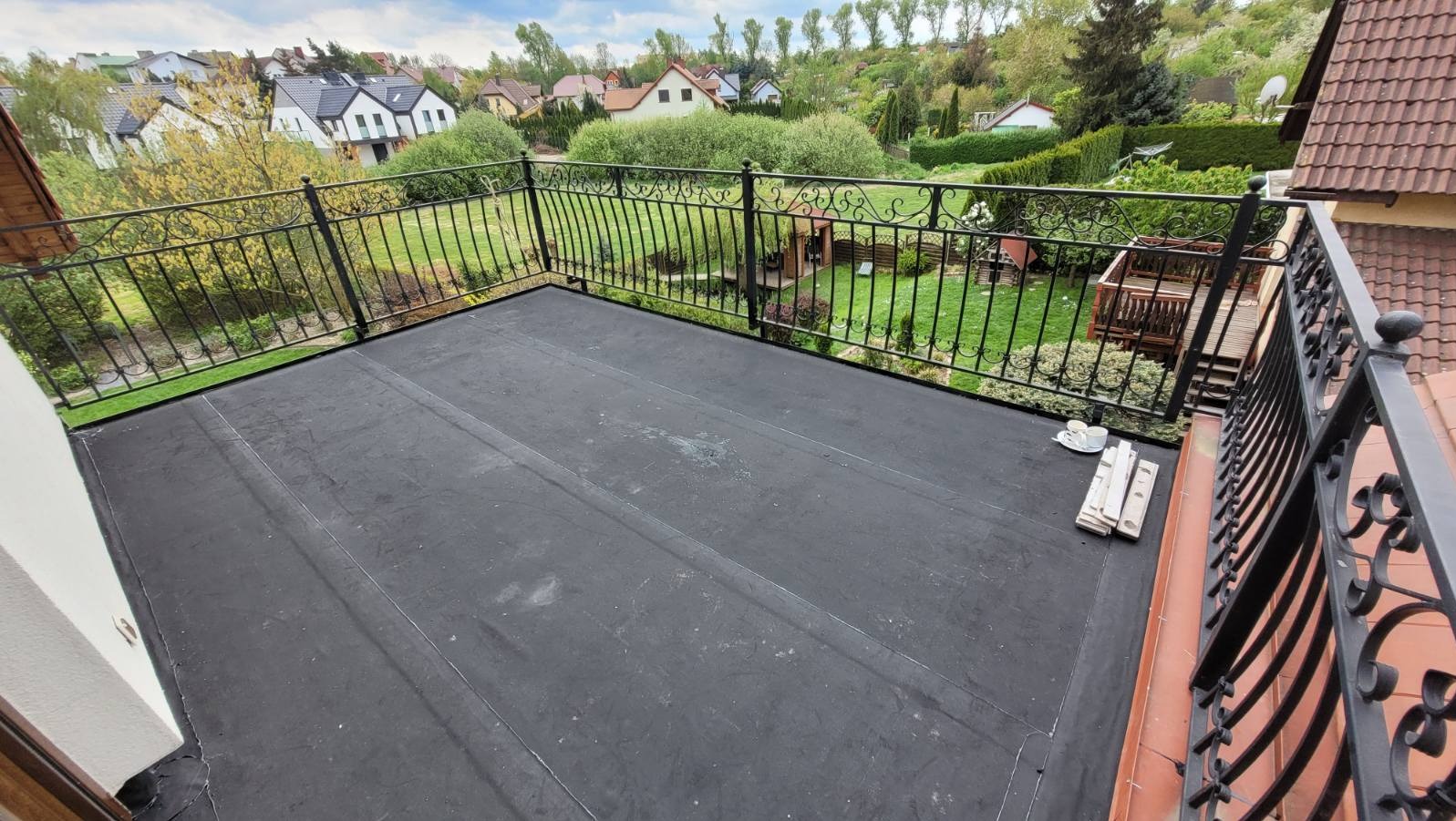 Wykończony płaski dach pokryty czarną papą termozgrzewalną w Poznaniu, z ozdobną, metalową balustradą i widokiem na zielony ogród oraz zabudowę jednorodzinną w tle. Na dachu leżą białe deski...