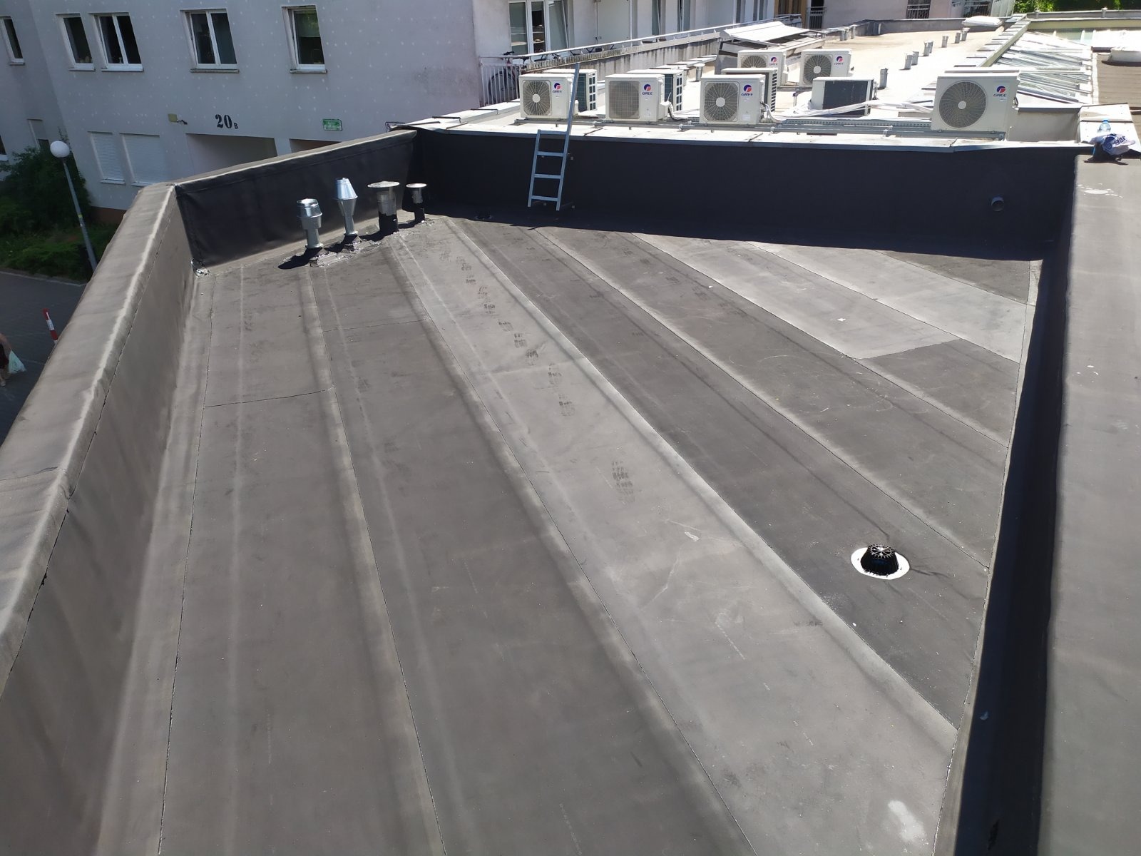 Czarny, płaski dach pokryty papą termozgrzewalną z widocznymi kominami wentylacyjnymi, wyłazem dachowym i licznymi klimatyzatorami na dachu budynku mieszkalnego w tle.