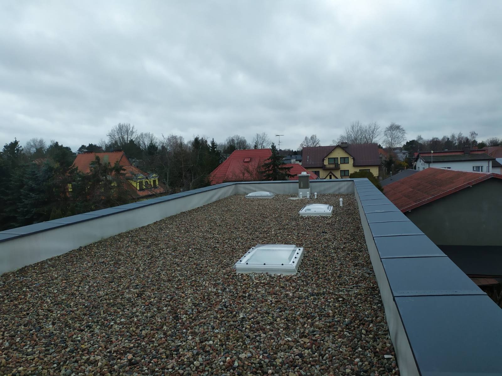 Płaski dach pokryty drobnym żwirem z trzema świetlikami i elementami wentylacyjnymi, widok z góry na tle zabudowy jednorodzinnej w pochmurny dzień.