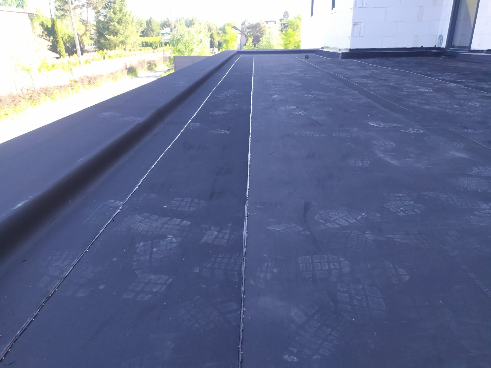 Czarna, płaska powierzchnia dachu pokryta papą termozgrzewalną z widocznymi śladami butów, z elementami konstrukcyjnymi i fragmentem budynku w tle.
