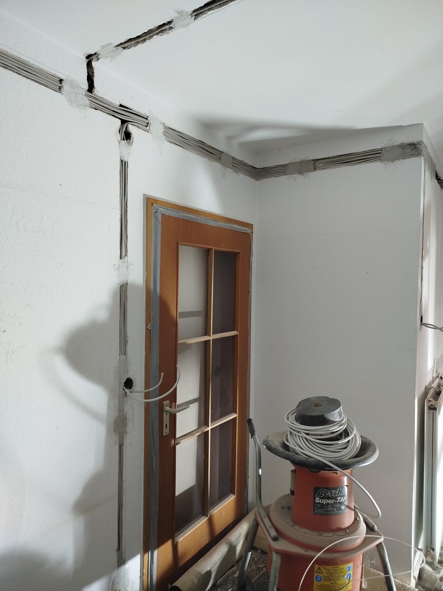Przygotowane podtynkowo przewody elektryczne w bruzdach na ścianie i suficie, częściowo zamaskowane gipsem, obok stoi odkurzacz przemysłowy z przewodem.