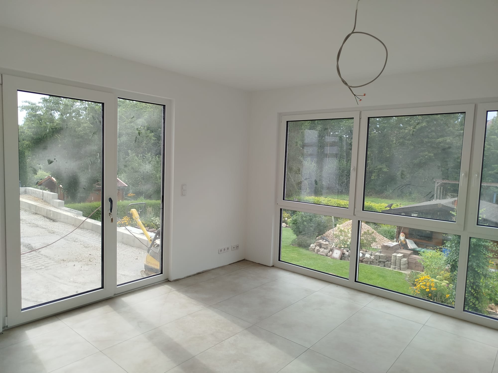 Jasne wnętrze z dużymi oknami i szarymi płytkami podłogowymi, widoczne wystające kable z sufitu, na zewnątrz krajobraz z ogrodem i ubijarką do gruntu.