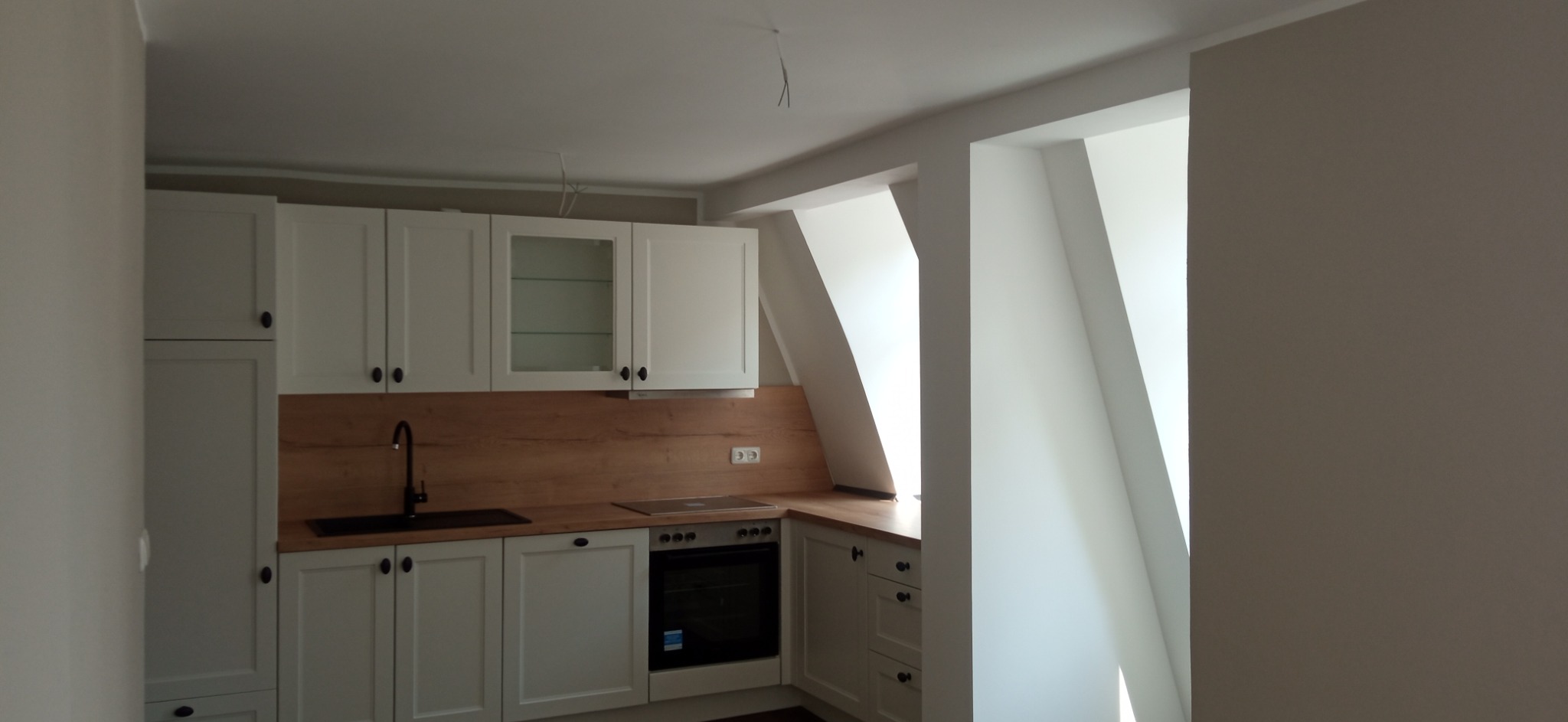 Jasna kuchnia w skosach poddasza z białymi szafkami, drewnianym blatem i czarną armaturą, widoczne gniazdka elektryczne i wystające przewody oświetleniowe.