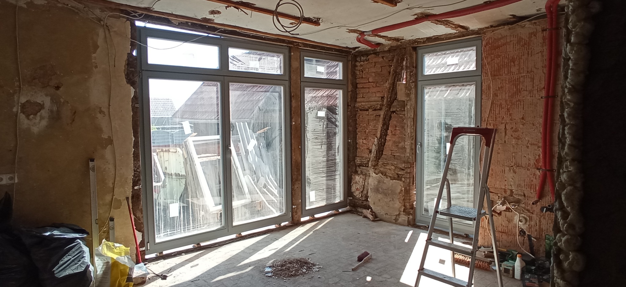 Wnętrze w trakcie remontu z trzema oknami z szarymi ramami, odsłoniętą cegłą, instalacją grzewczą z czerwonych rur na suficie i drabiną.