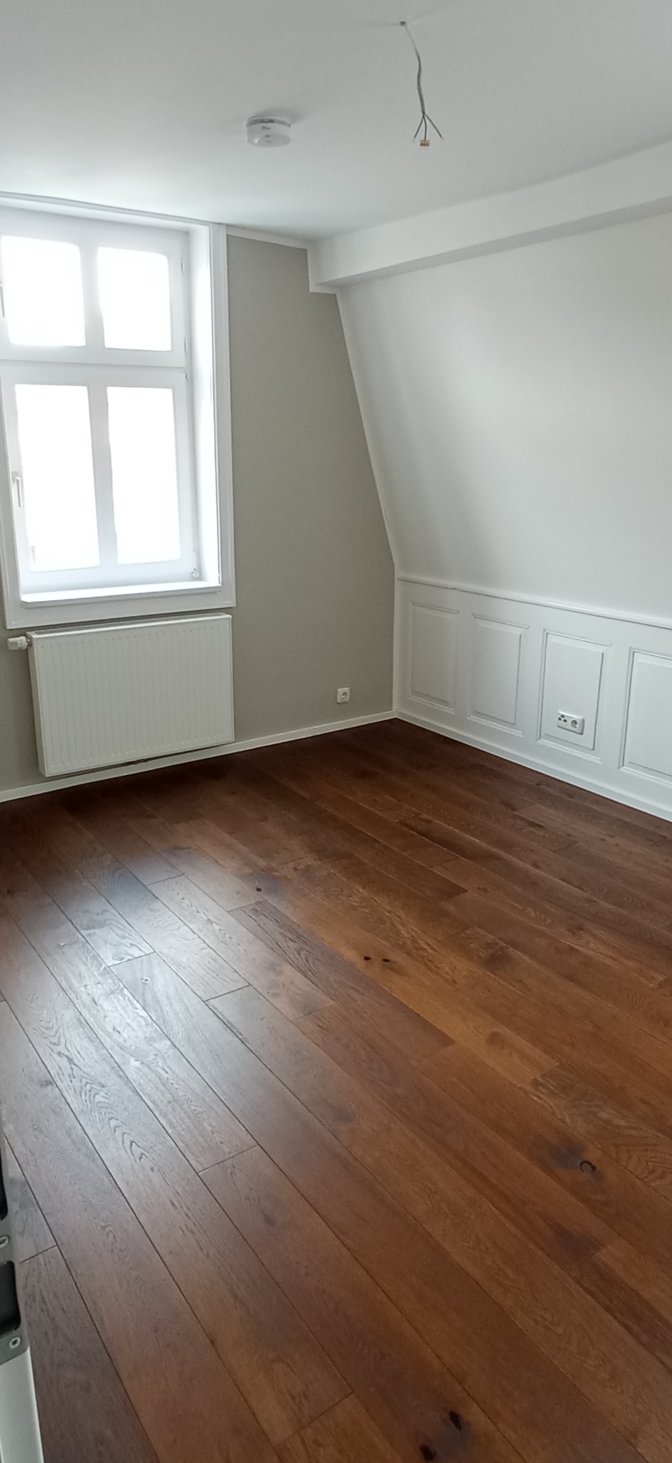 Pusty pokój po remoncie z drewnianą podłogą, szarą ścianą z oknem i białymi panelami ściennymi. Widoczne gniazdka elektryczne i wystające przewody z sufitu.