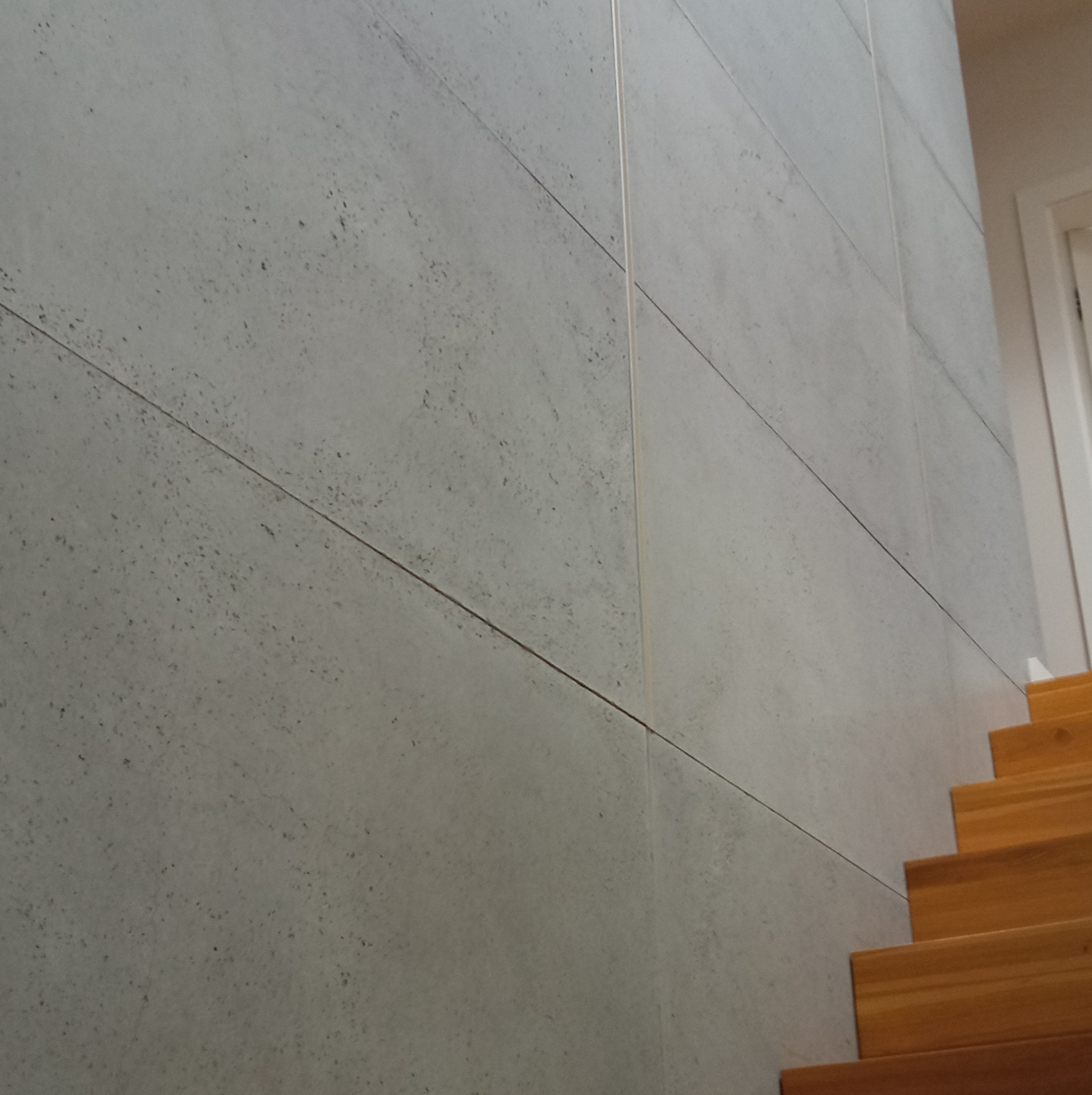 Ściana wykończona szarymi panelami imitującymi beton, z widocznymi drewnianymi schodami po prawej stronie, ujęcie z dołu.