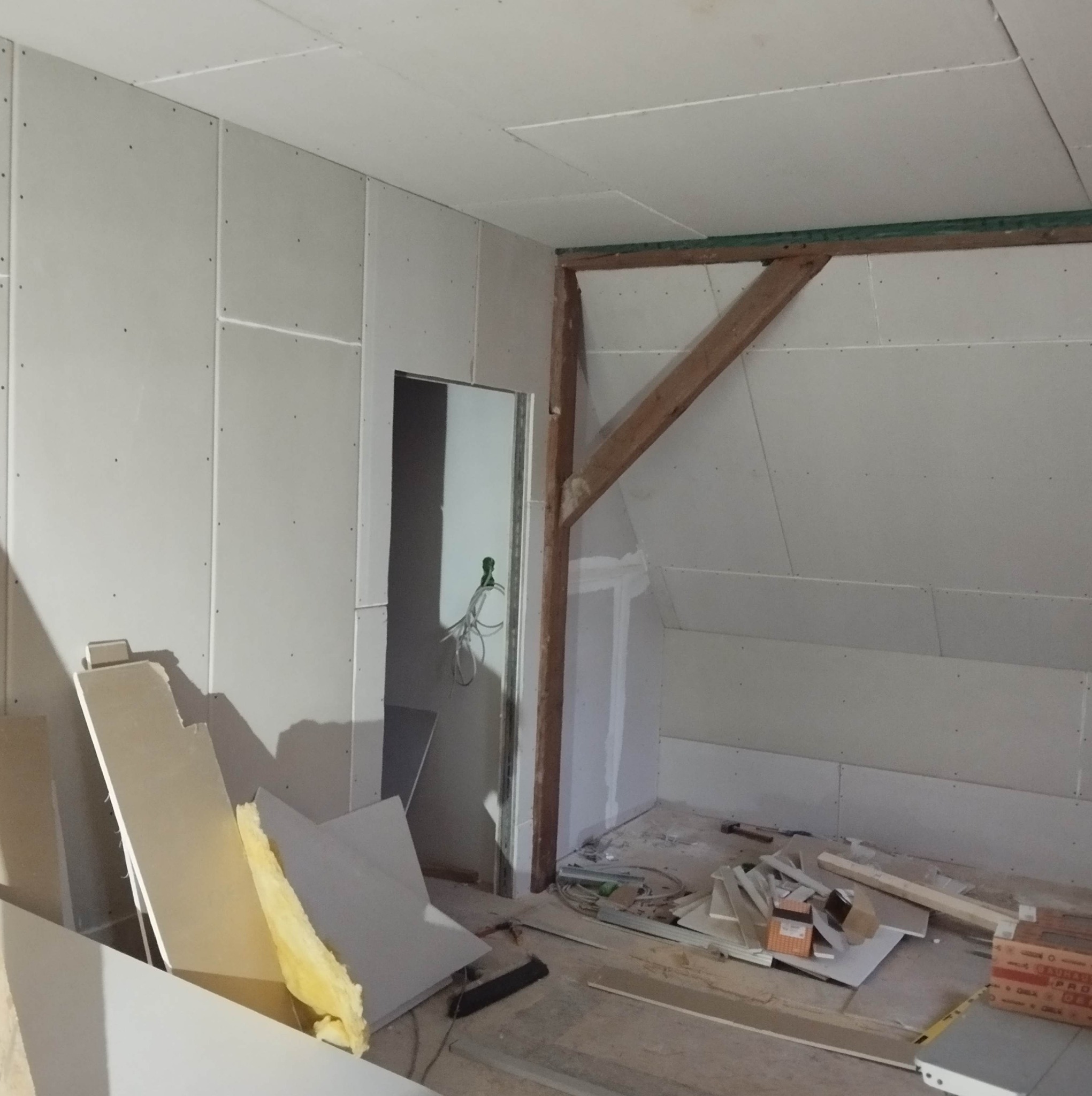 Poddasze w trakcie wykańczania, ściany i sufit pokryte płytami gipsowo-kartonowymi, widoczne drewniane elementy konstrukcyjne dachu, na podłodze resztki materiałów budowlanych i izolacji.